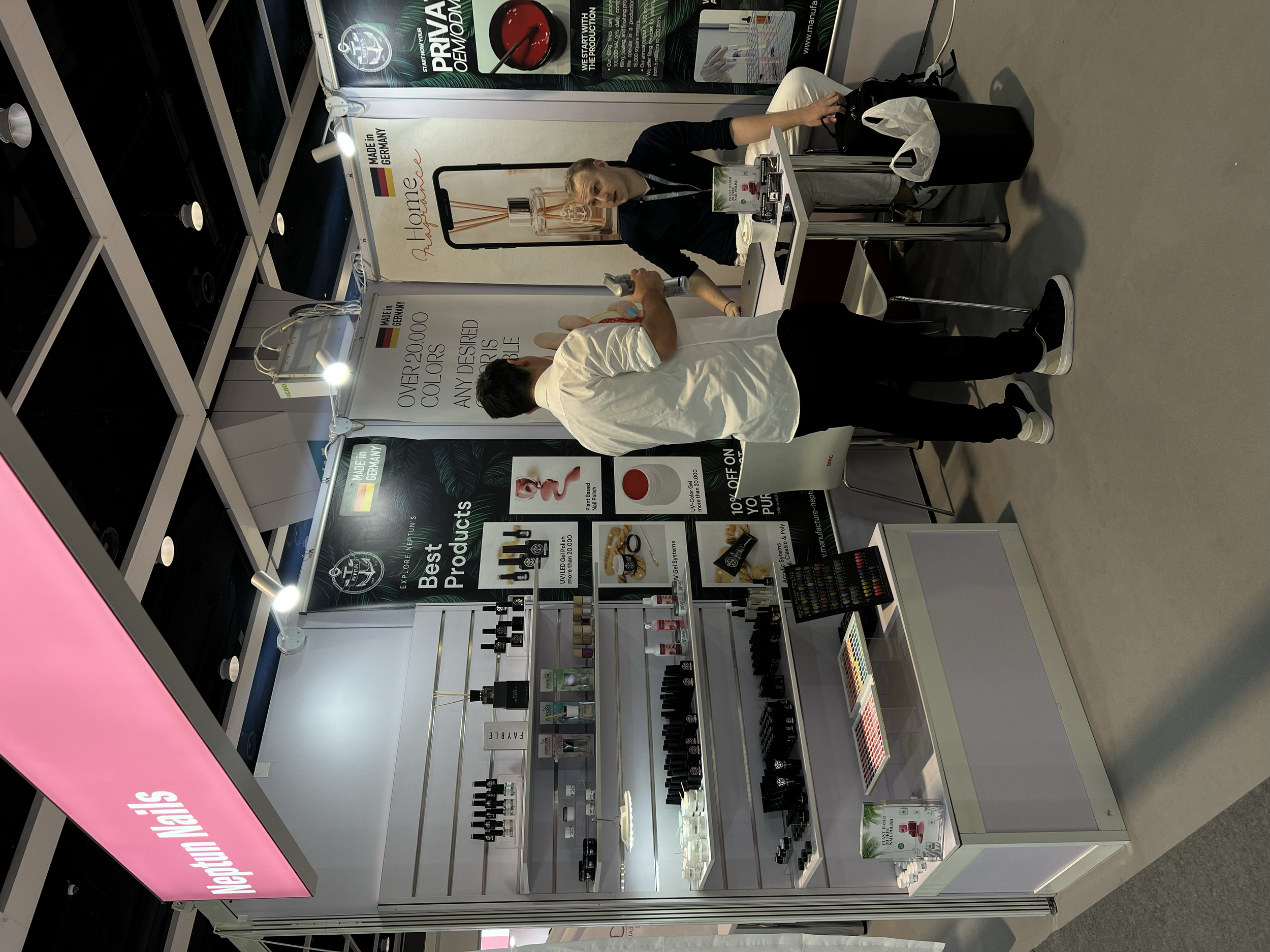 A display booth at a trade show selling nail polish and cosmetics, with a pink sign reading 'Nail Polish Nails.' There is a man talking to a woman behind the booth, which has various nail polish bottles, makeup products, and promotional signs. The wo