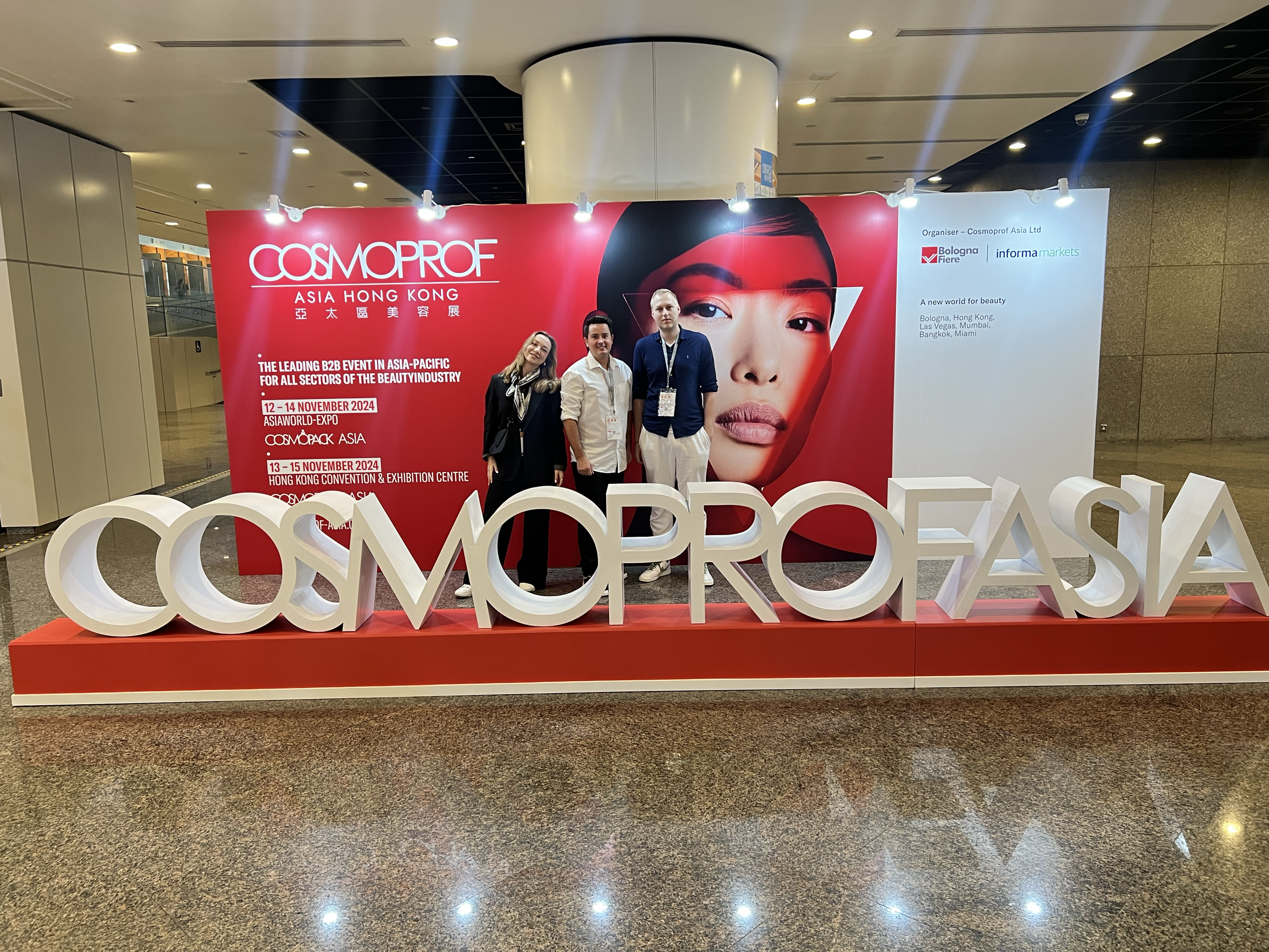 Photograph of three people standing in front of a red and white promotional display for Cosmoprof Asia Hong Kong. The display features a large image of a woman's face and text about the event, with the large 3D sign of 'COSMOPROFASIA' in front.