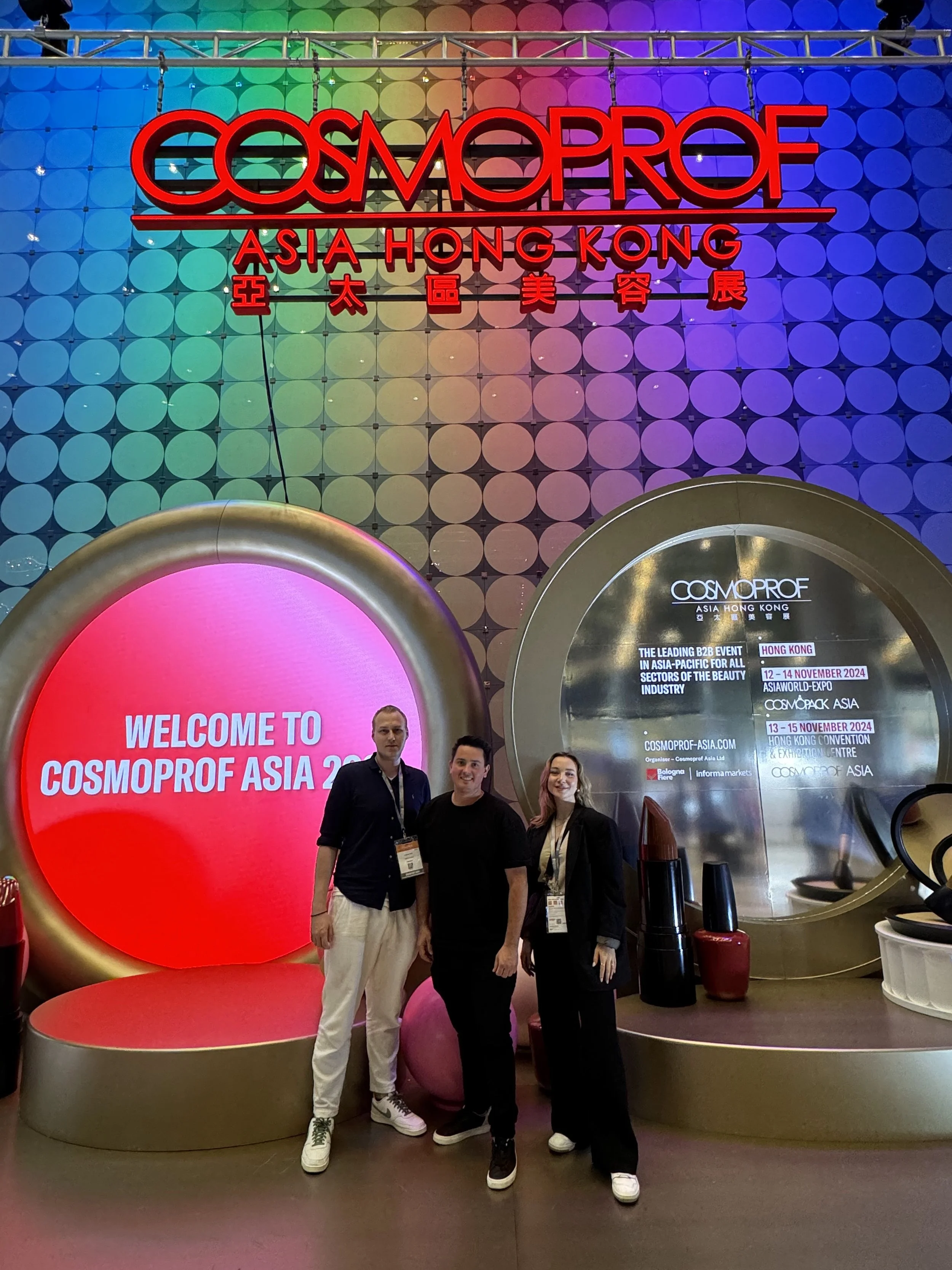 Three people standing in front of a colorful display at Cosmoprof Asia Hong Kong 2024. The background includes a large, illuminated sign with the event name and dates, and a screen that says 'Welcome to Cosmoprof Asia 2024.' The display features beauty products like lipstick and nail polish.