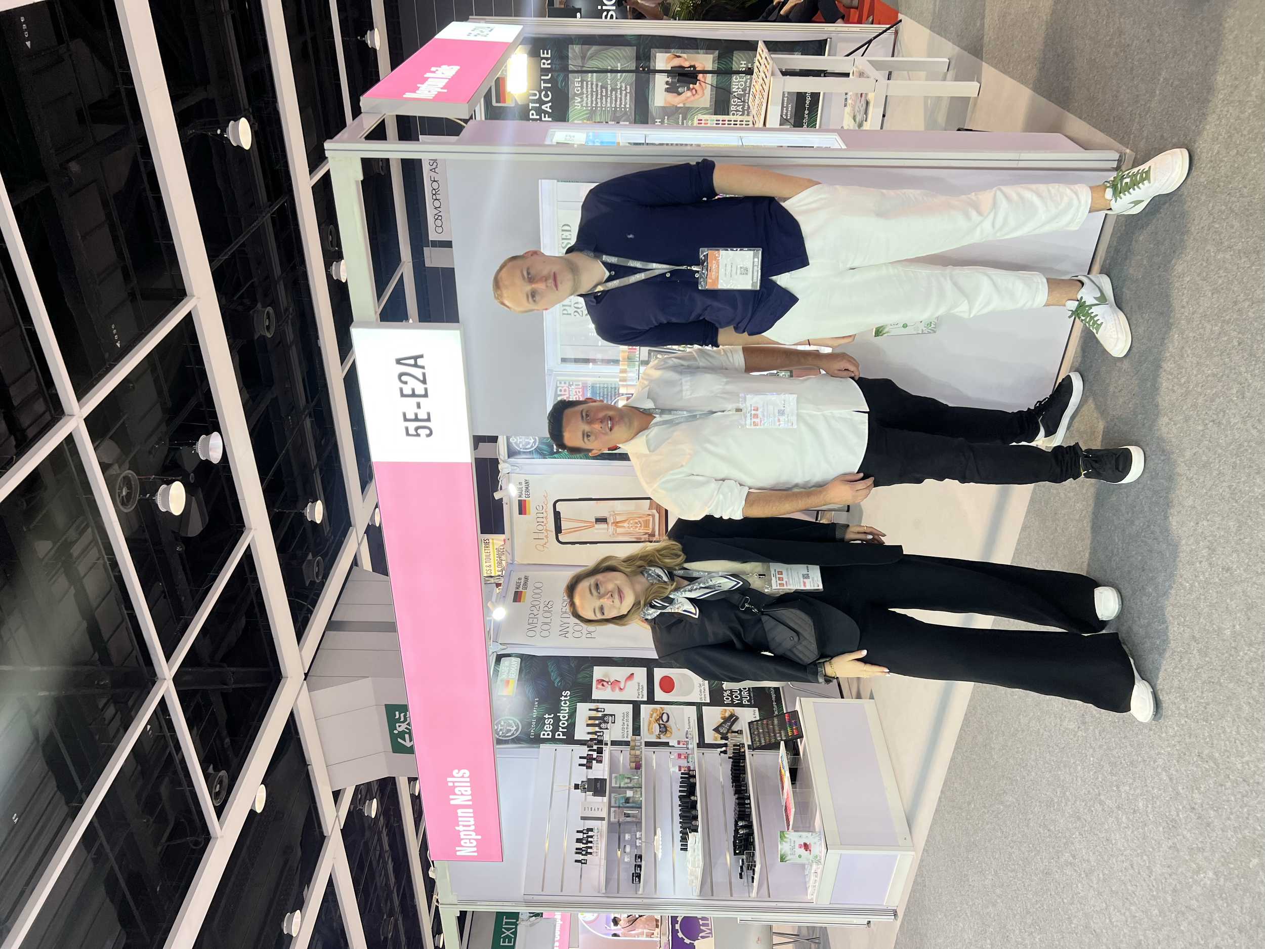 Three people standing in front of a beauty product booth at a trade show, with shelves displaying nail care products. They are wearing badges and smiling at the camera.