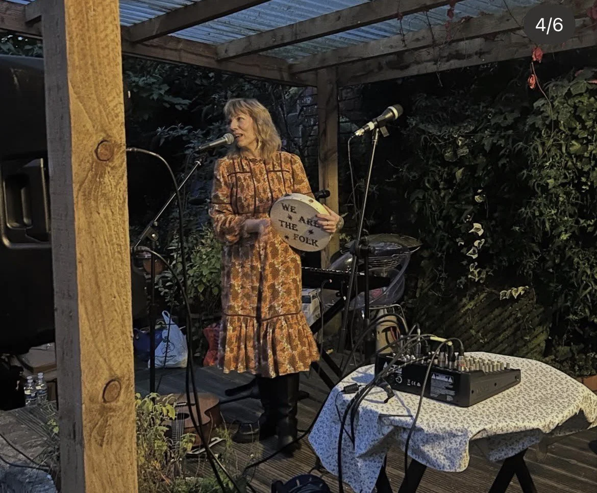 Woman in a floral dress holding a sign that says "We Are The Folk" standing on an outdoor wooden stage with microphones and sound equipment, surrounded by greenery and a wooden structure.