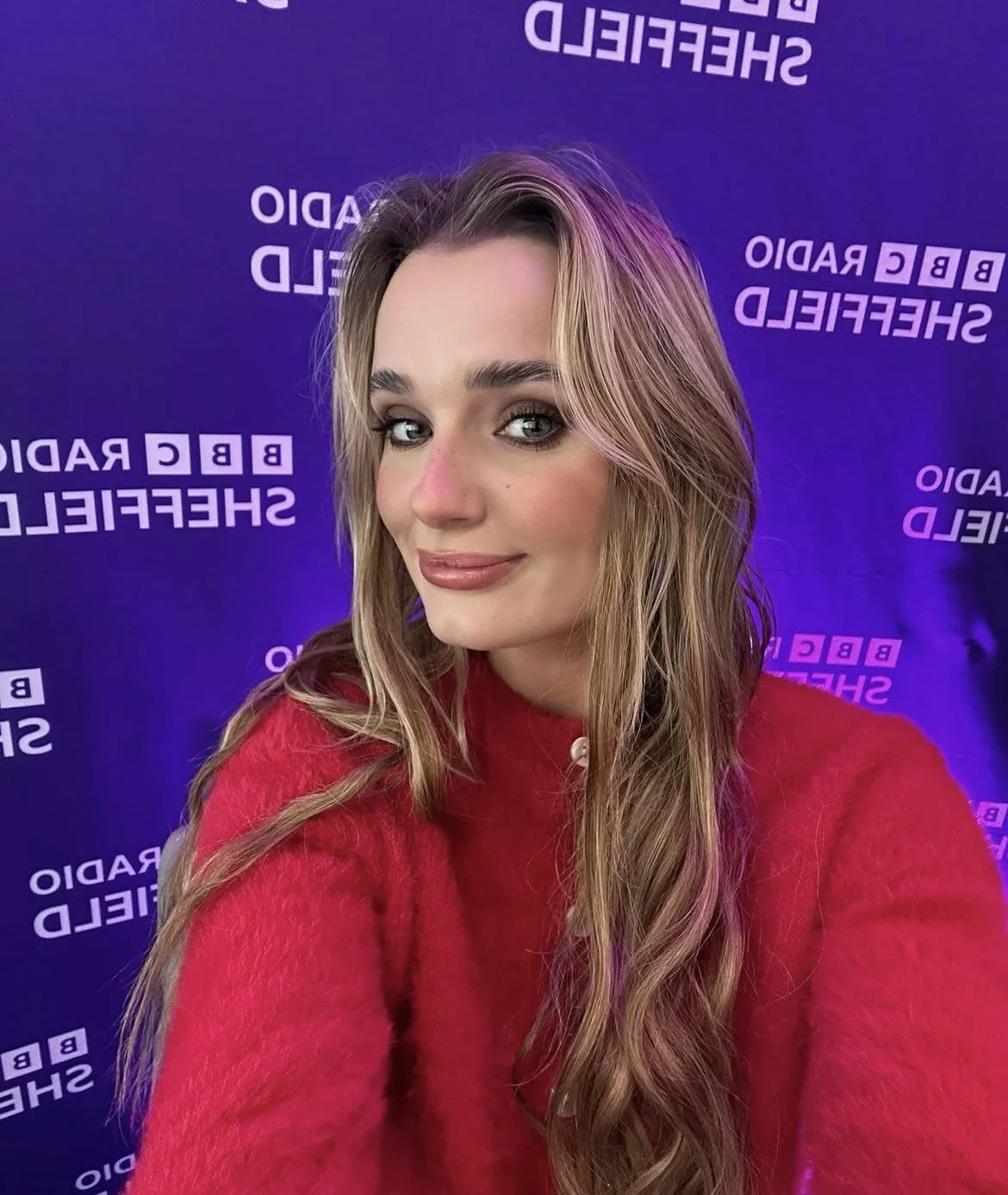 A woman with long wavy blonde hair taking a selfie, wearing a red sweater, in front of a purple backdrop with the logos 'BBC Radio Sheffield'.