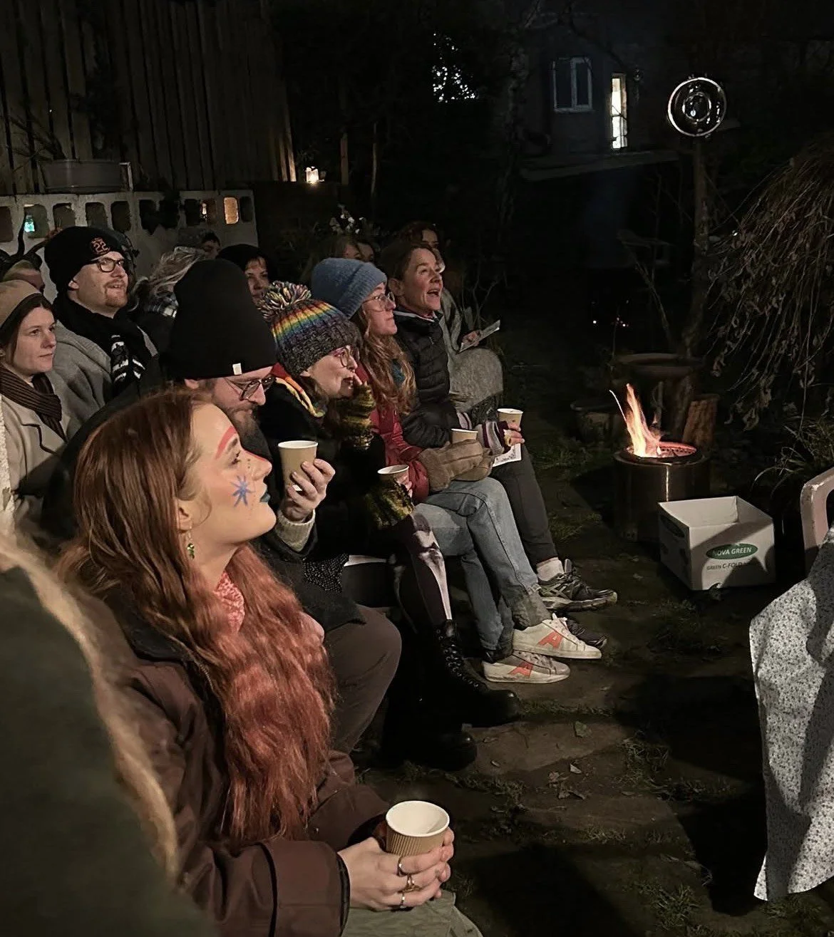 Group of people sitting outdoors at night around a small fire, watching a projection on a screen.