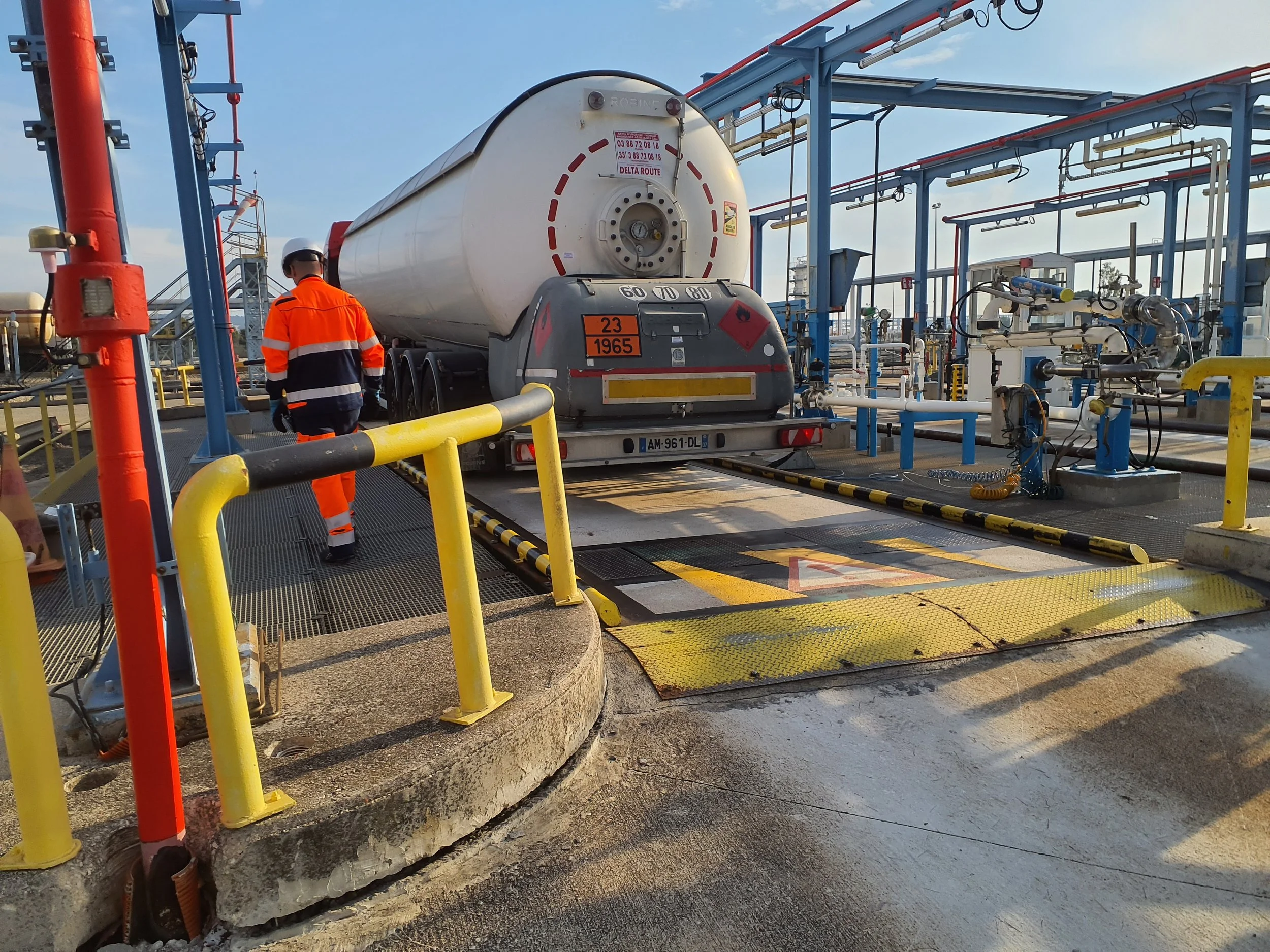 Un conducteur en tenue orange et casque se trouve à côté d’un camion-citerne stationné sur une plateforme de déchargement de produits chimiques, avec des tuyaux et des équipements industriels autour.