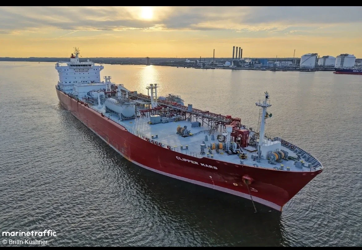 Un grand navire cargo rouge nommé 'Clipper Mars' navigue sur l'eau lors d'un coucher de soleil, avec des installations industrielles en arrière-plan.