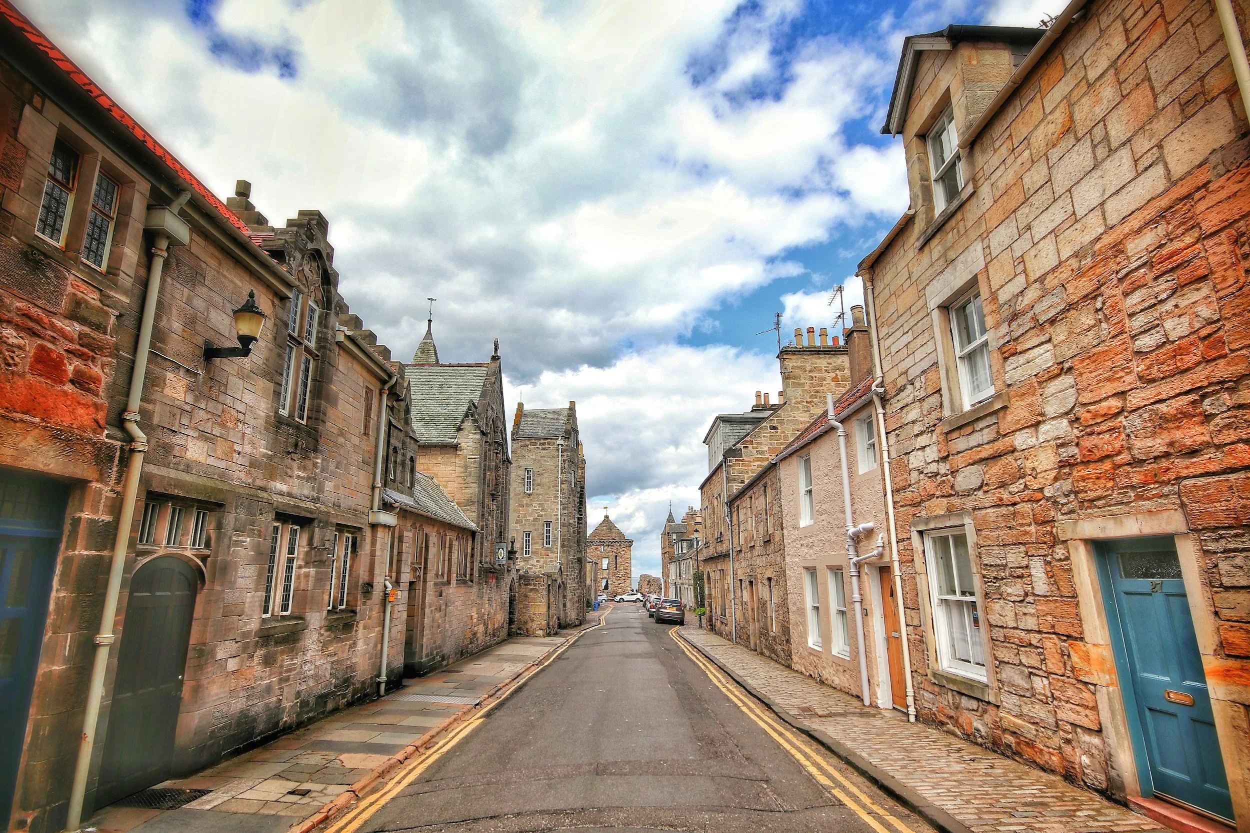A narrow street in a historic town with stone buildings, some with colorful doors, and a cloudy sky overhead.