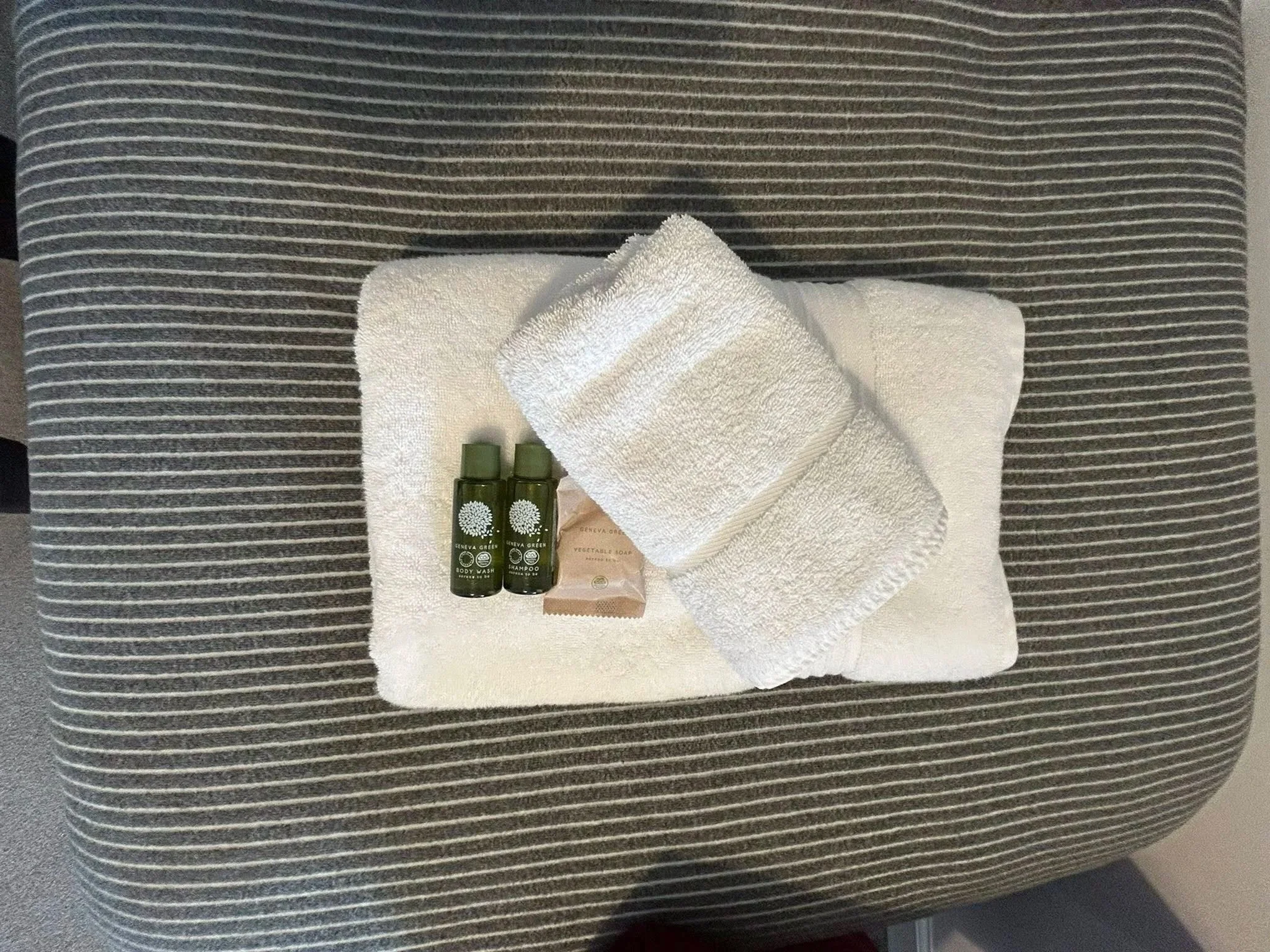 A set of hotel toiletries including two small green bottles, a bar of soap, and two white towels on a white cloth on a striped gray surface.
