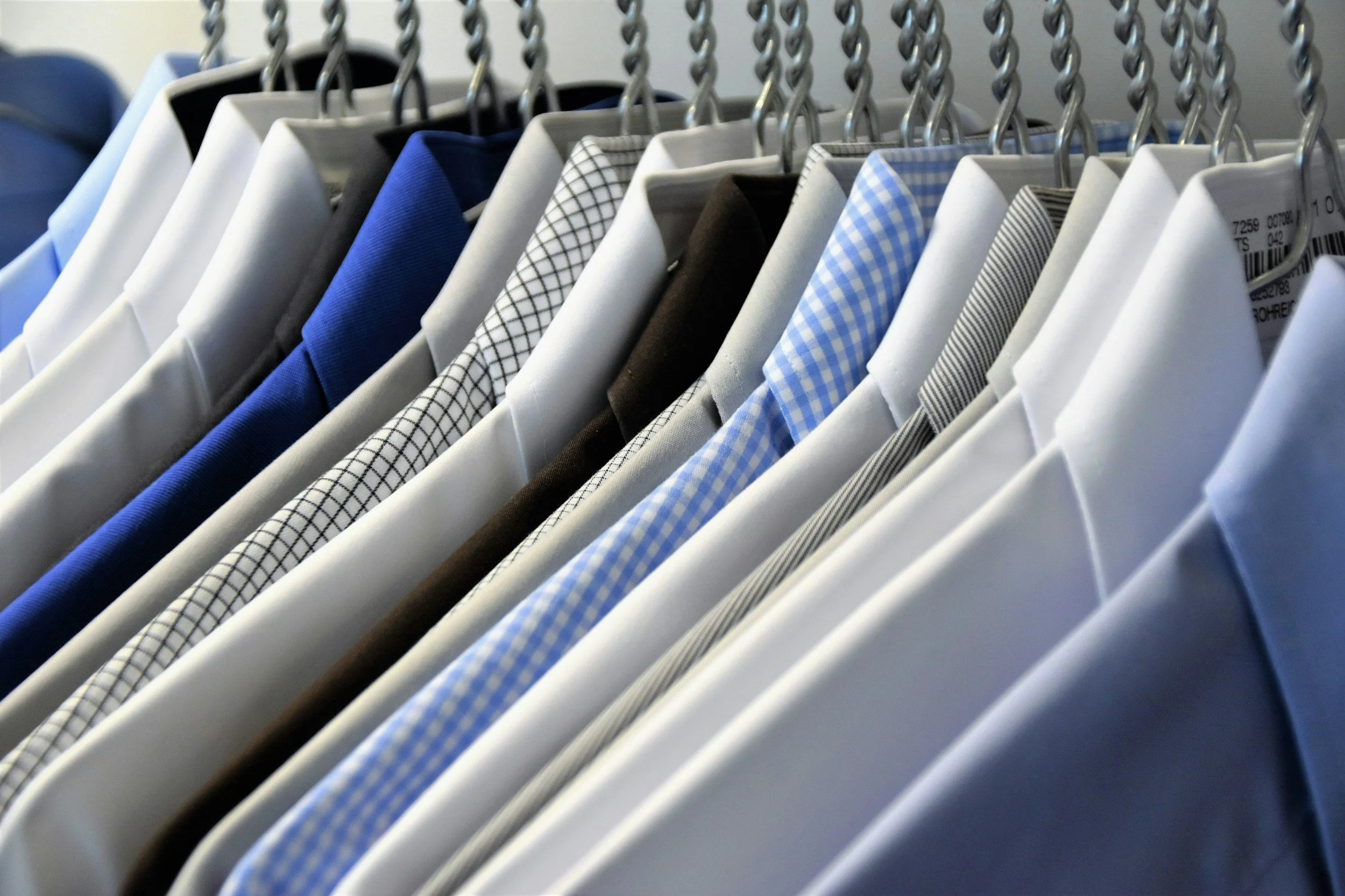 A row of men's dress shirts hanging on a clothing rack, featuring various colors and patterns including white, blue, black, and checkered designs.