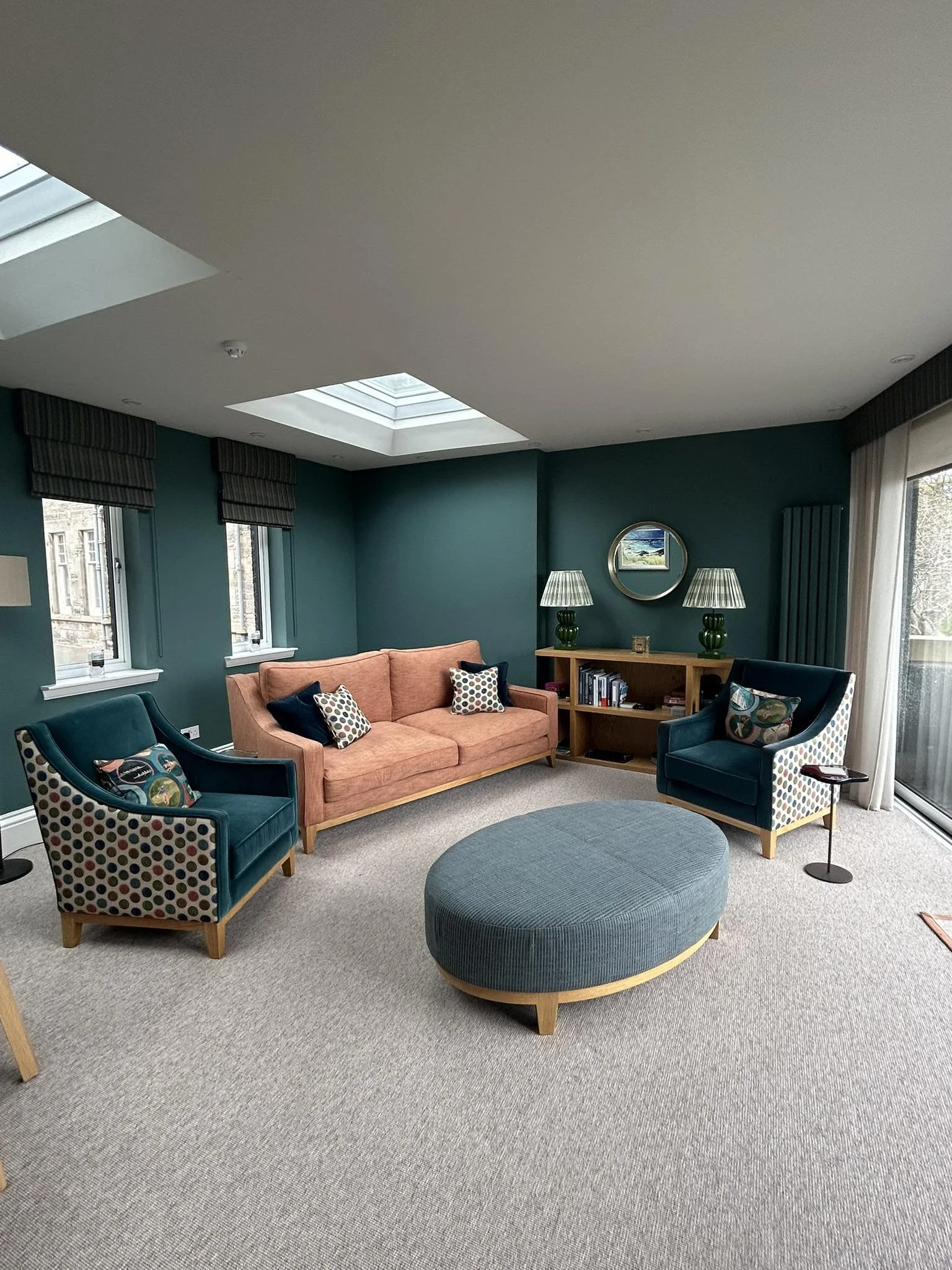 Living room with teal walls, large windows, beige carpet, pink sofa, two teal armchairs with polka dot pillows, round teal ottoman, wooden bookshelf with books, two green lamps with striped shades, round mirror, framed landscape picture, curtains, and ceiling skylights.