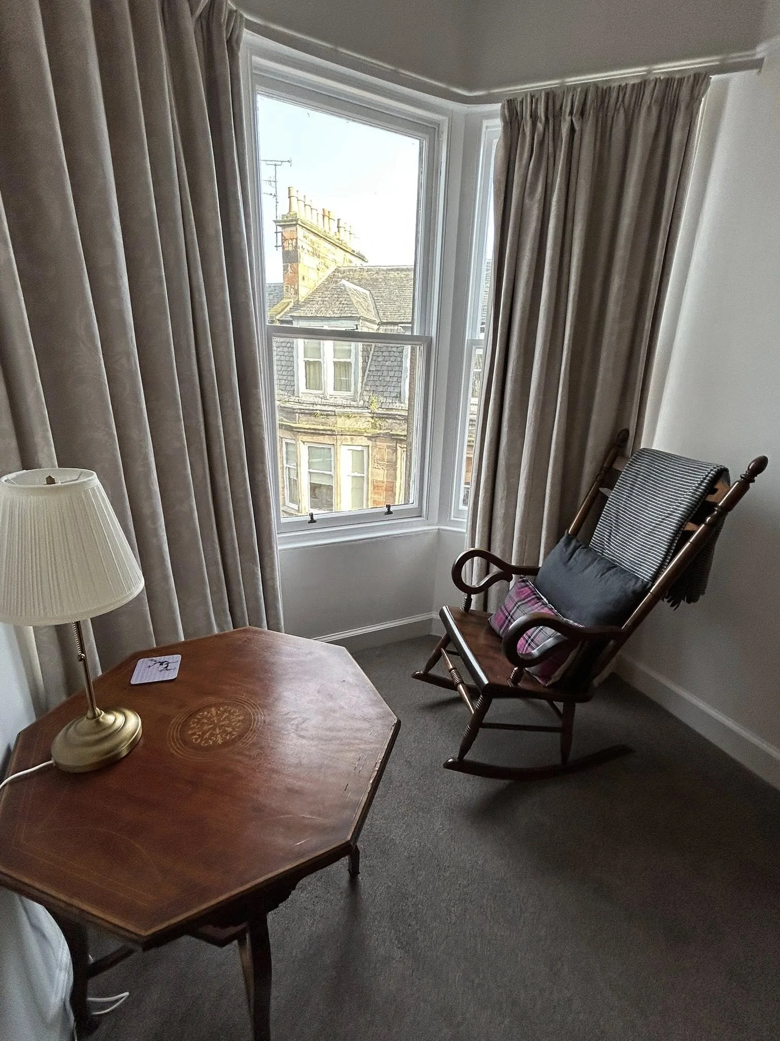 A cozy corner of a room with a wooden rocking chair with cushions, a small wooden table, a table lamp, and a large window with curtains showing a view of neighboring buildings.