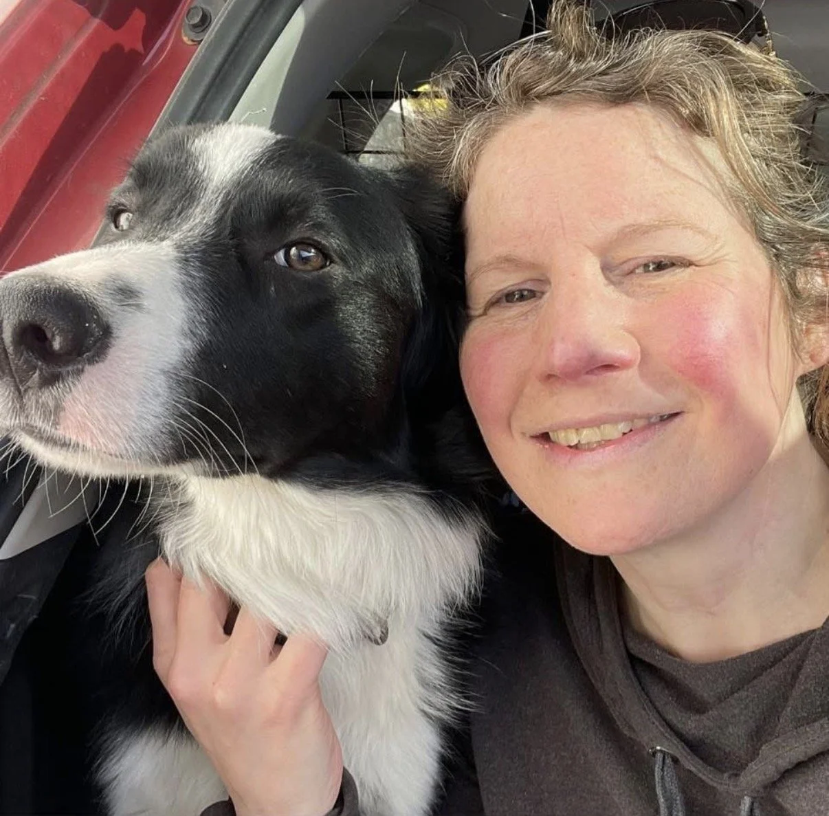 A woman with short, light brown hair and a black zip-up jacket smiling while taking a selfie with a black and white dog, possibly a Border Collie mix, sitting beside her in a vehicle.