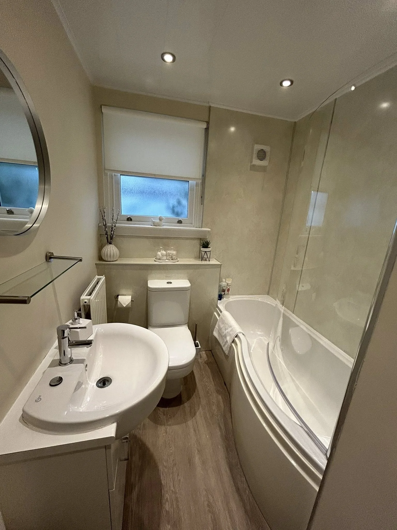 Small modern bathroom with a white sink, toilet, bathtub with a glass screen, window with shades, a round mirror, and decorative items on the window ledge.