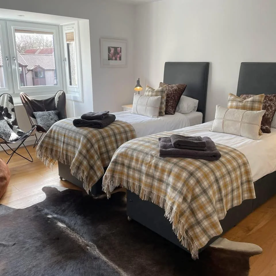 Two beds with plaid blankets and multiple pillows in a bedroom with wooden floor, black cowhide rug, and chair near a window.