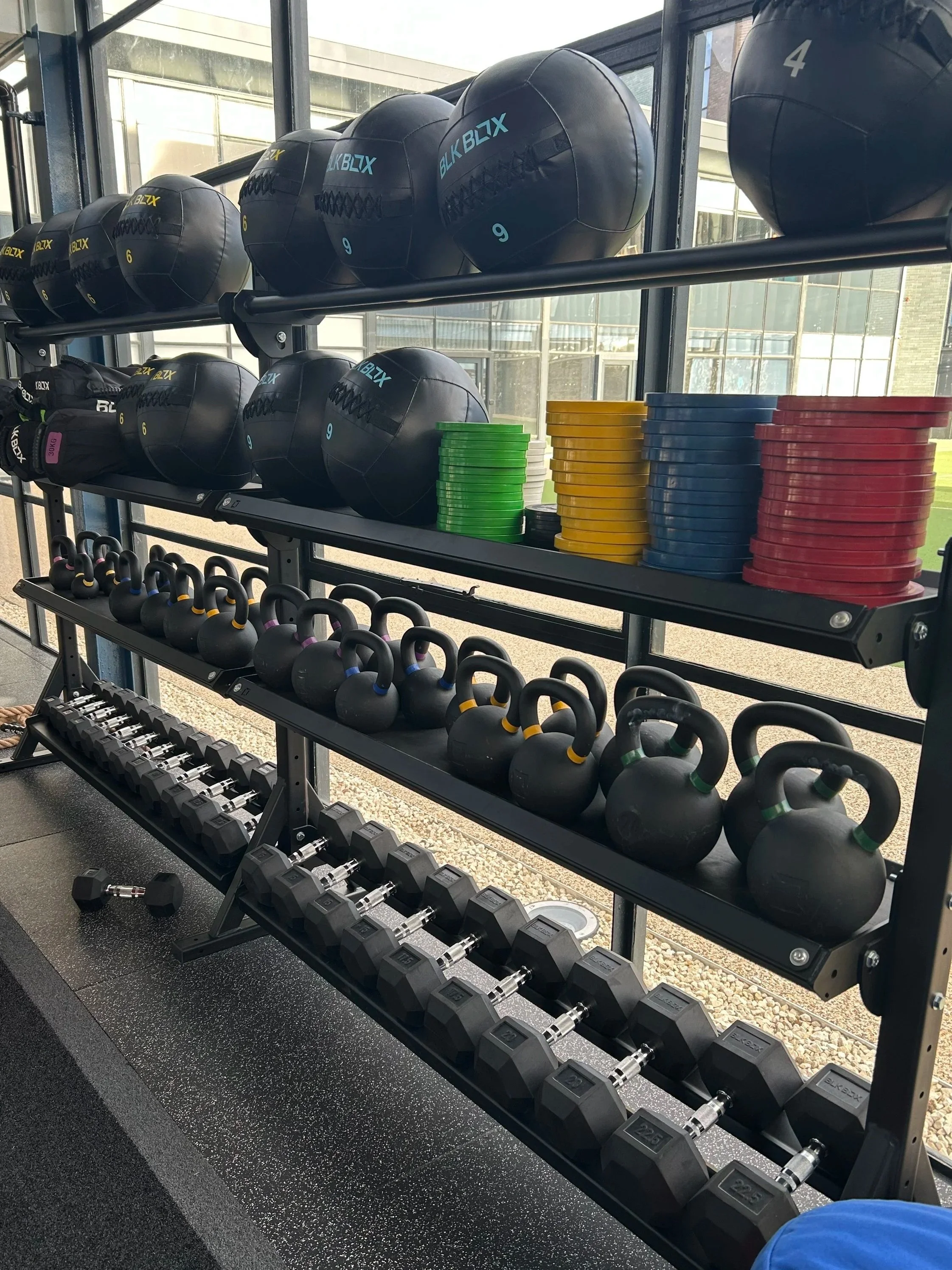 Rack of black medicine balls, colored weight plates, kettlebells, and dumbbells in a gym with large windows.