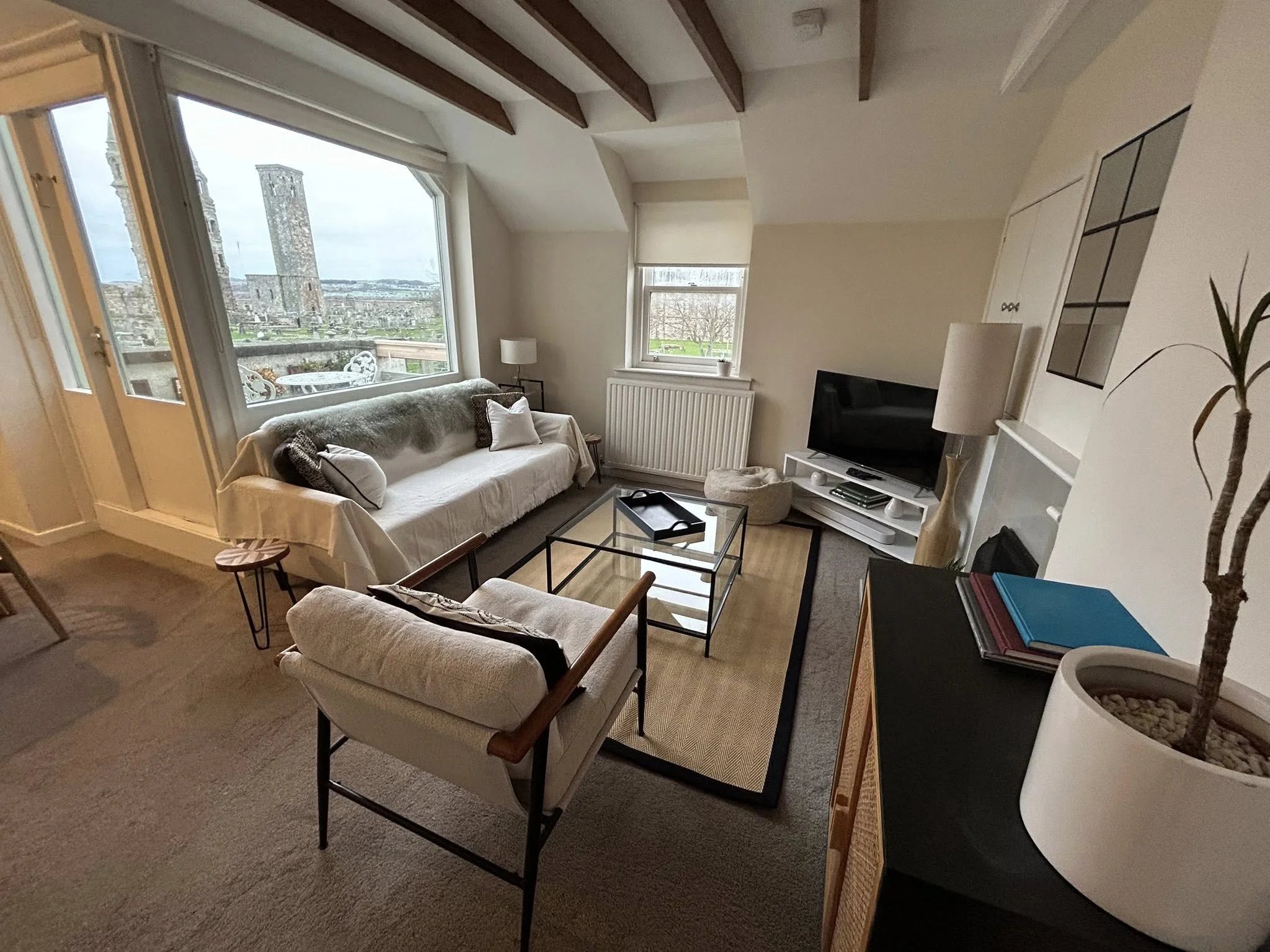 Living room with a large window overlooking a cityscape, white sofa with cushions, glass coffee table, armchair, television, bookshelf, and decorative items.