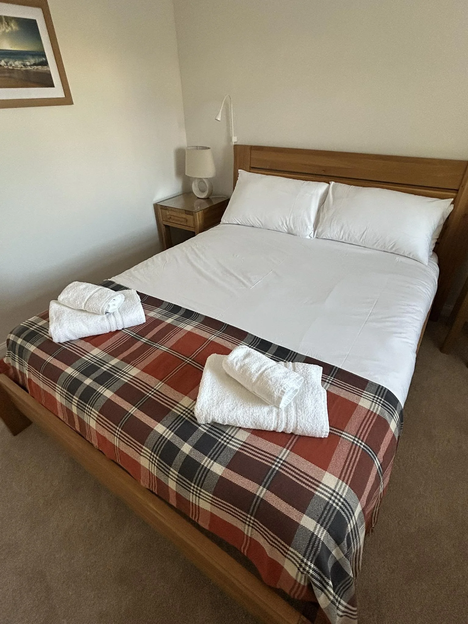 A neatly made bed with white sheets, pillows, and a plaid blanket at the foot, topped with rolled white towels, in a hotel room with a wooden nightstand, table lamp, and artwork on the wall.