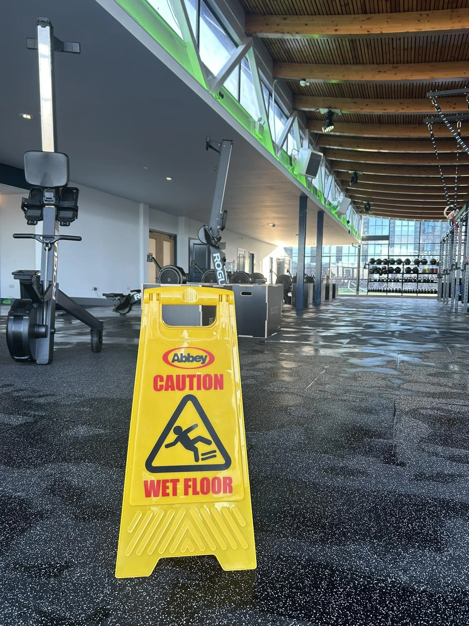 Wet floor caution sign in a gym with workout equipment and a high ceiling with wooden beams.