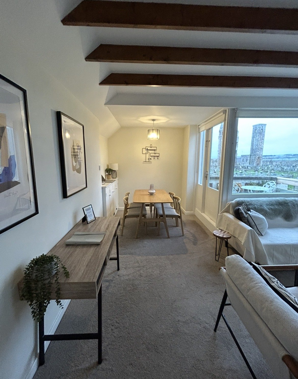 Living and dining room with large windows view of cityscape, wooden ceiling beams, white walls, beige carpet, modern furniture, and wall art.