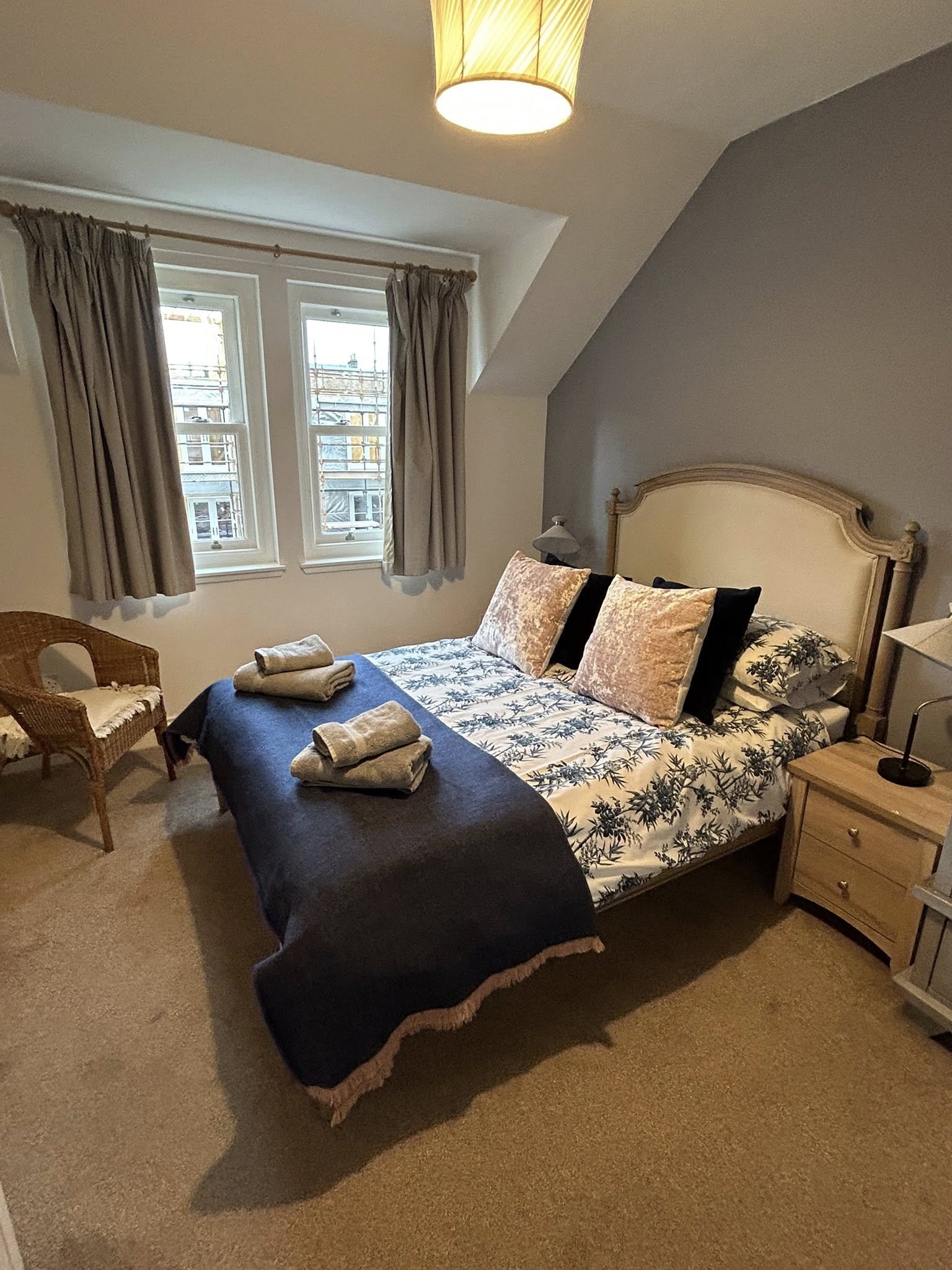 A cozy bedroom with a bed covered in floral bedding and decorative pillows, beside a wooden nightstand with a lamp, a wicker chair, and double windows with beige curtains, during daytime.