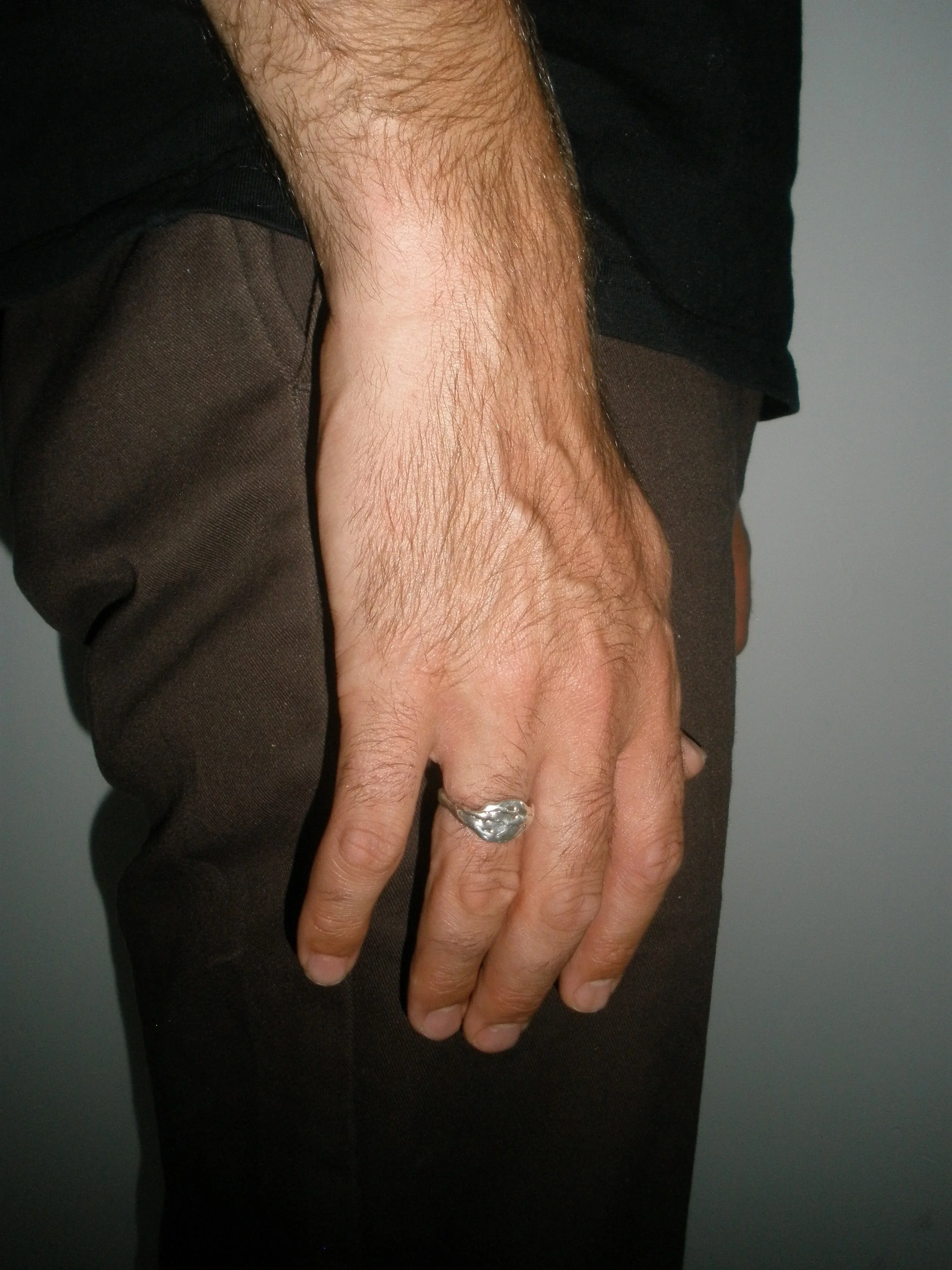 A close-up of a person's hand with a silver ring on the middle finger, wearing a black shirt and brown pants, against a grey background.