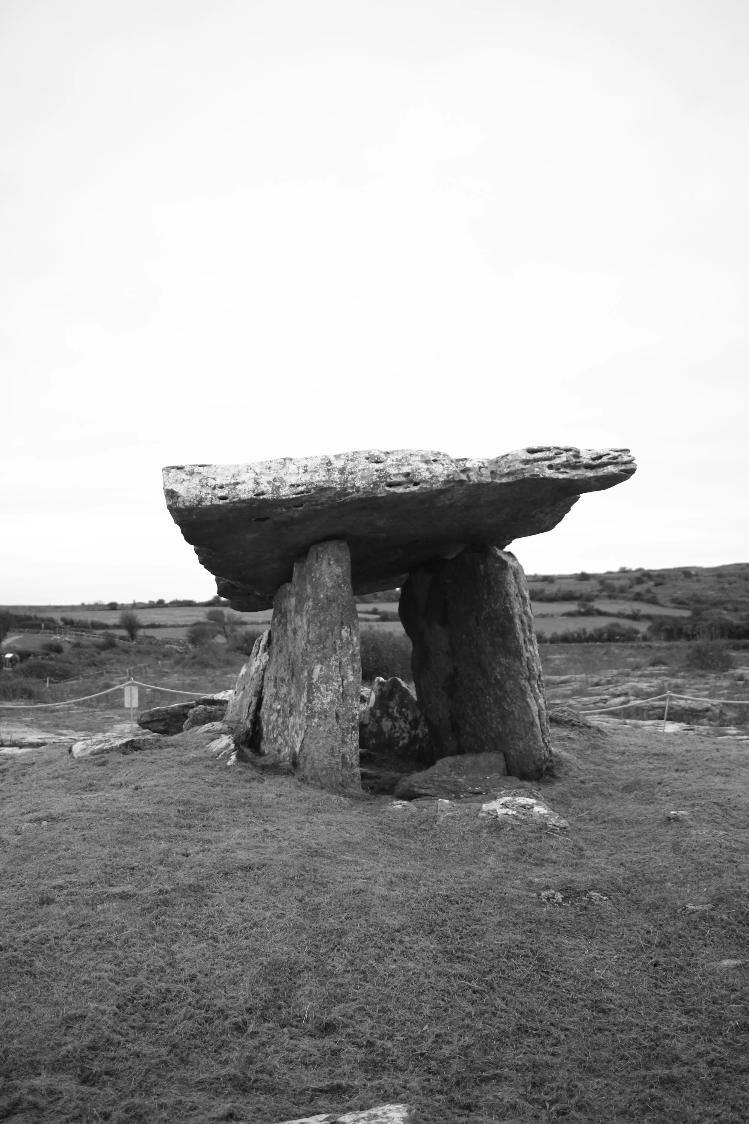 Black and white photo of a stone dolmen with large upright stones supporting a flat capstone, situated on a grassy landscape under an overcast sky.