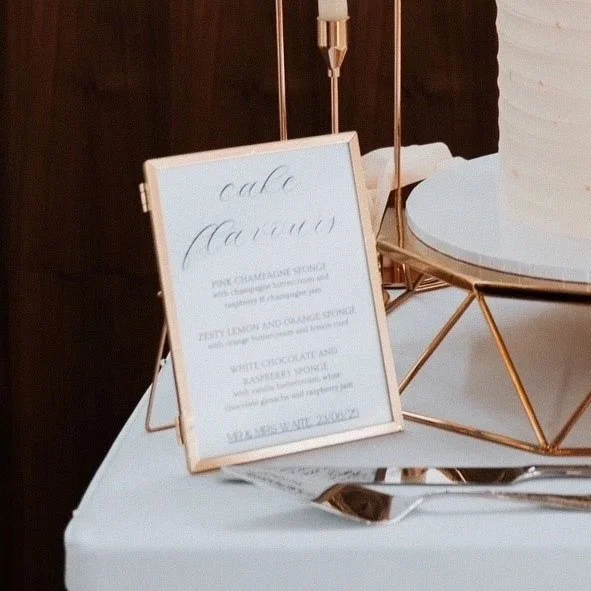 A whiteboard menu with handwritten lettering listing dessert options, placed on a table with a serving spoon, a white cake on a geometric stand, and a small card, in a decorative setting.