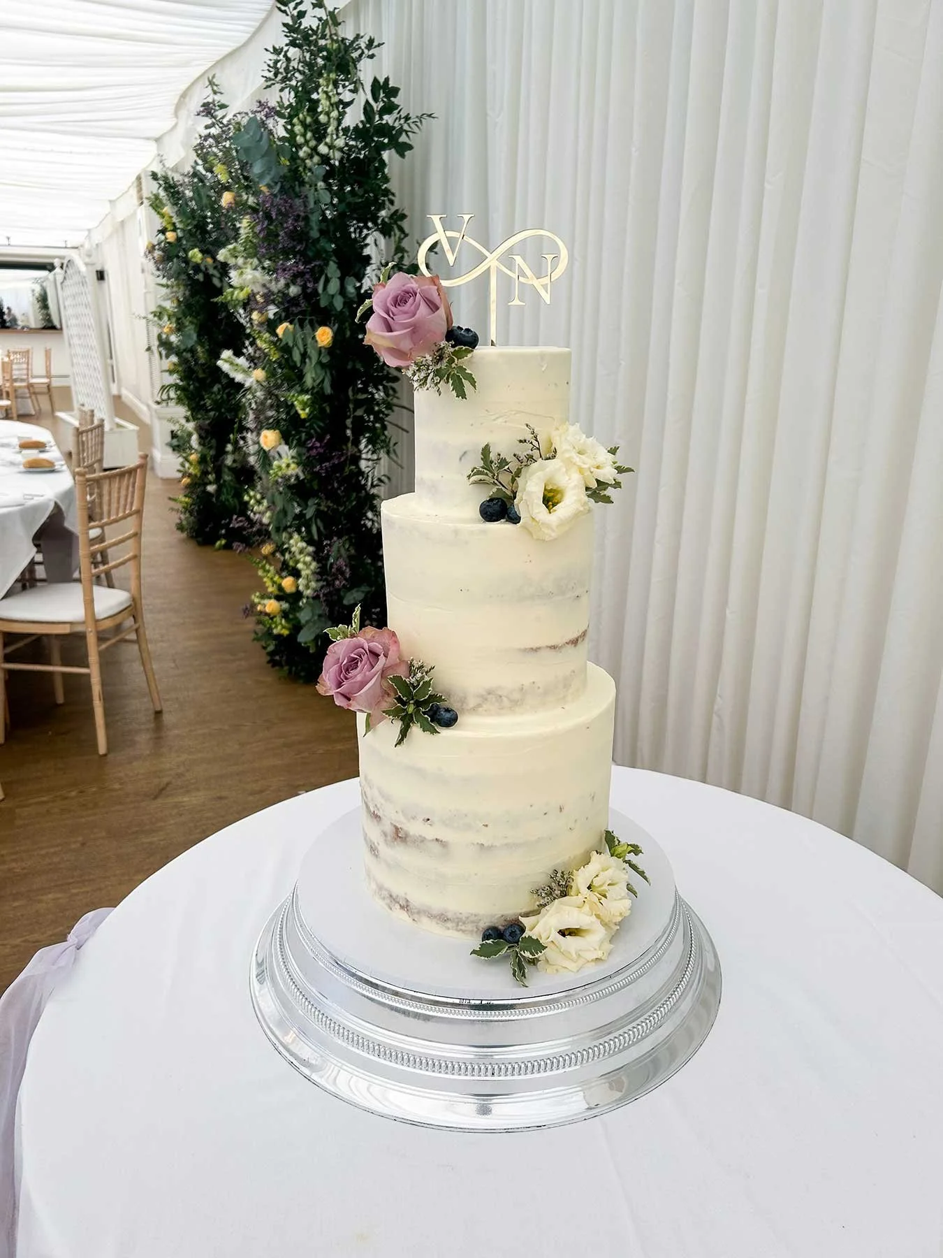 Three-tiered wedding cake decorated with pink roses, white flowers, blueberries, and greenery, topped with a gold Infinity monogram cake topper, placed on a silver stand in an event hall.