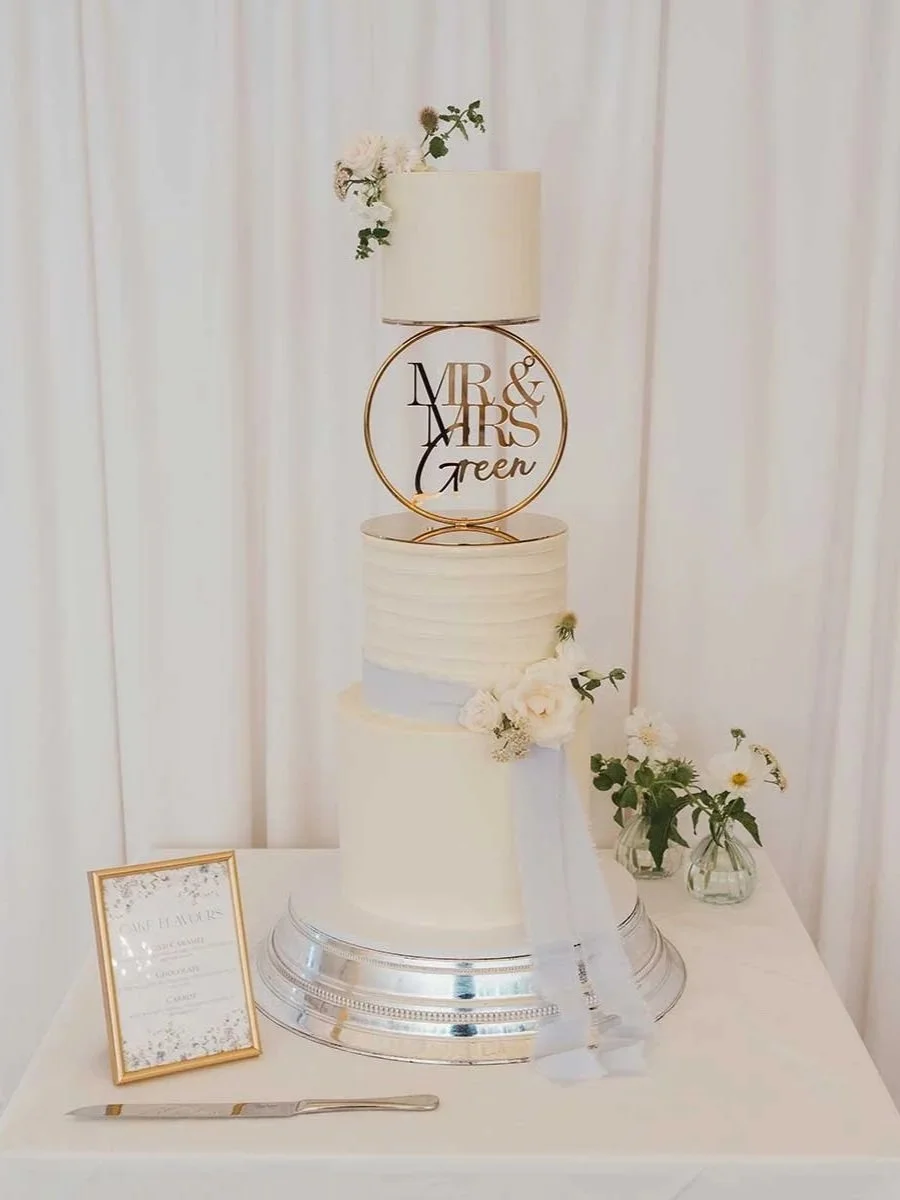 Wedding cake with two tiers decorated with white flowers and greenery, topped with a gold "Mr & Mrs Green" cake topper, on a silver stand. Behind the cake are two small vases with white flowers, and a framed menu is on the table.