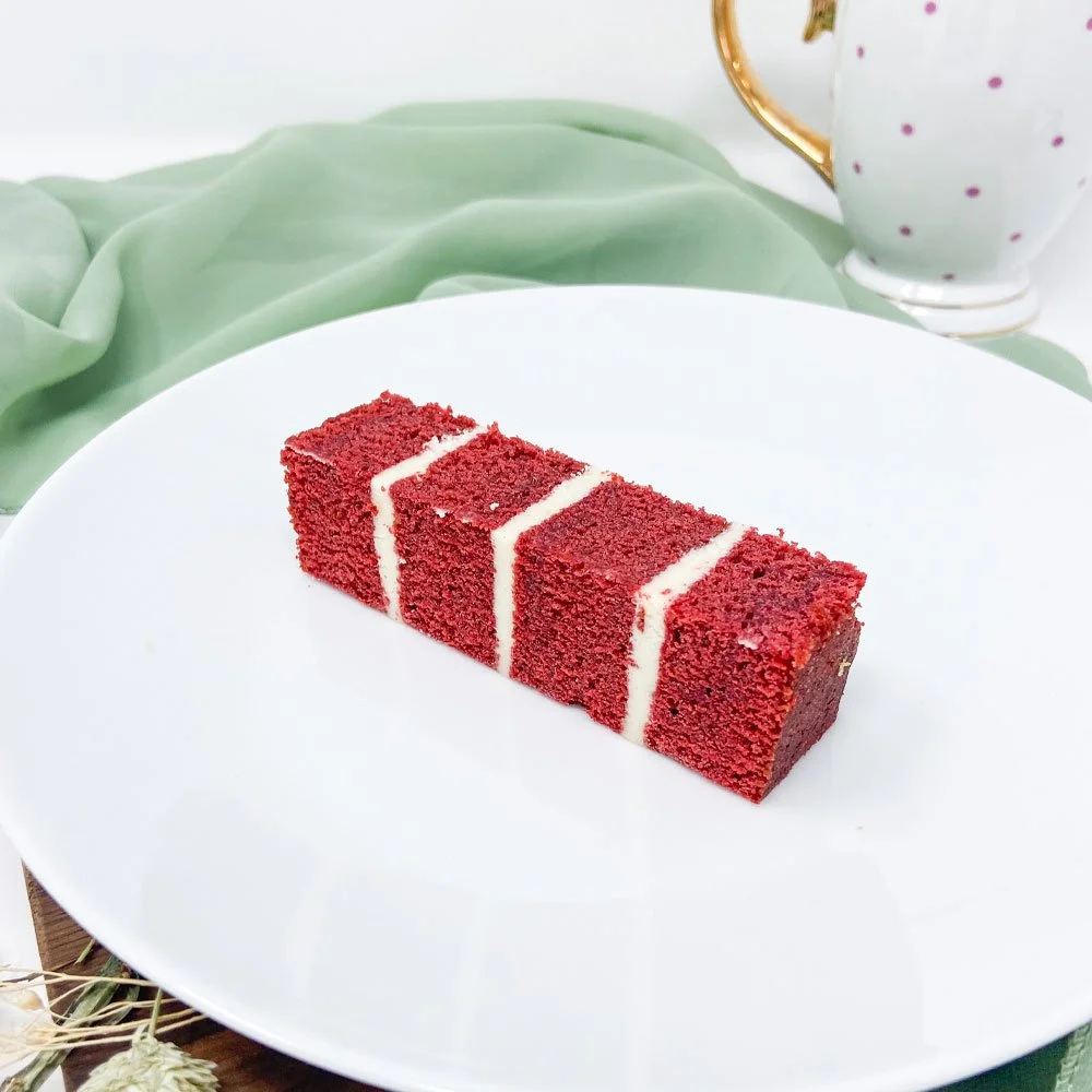 A slice of red velvet cake with cream cheese frosting on a white plate, with a polka dot teacup and a green cloth in the background.
