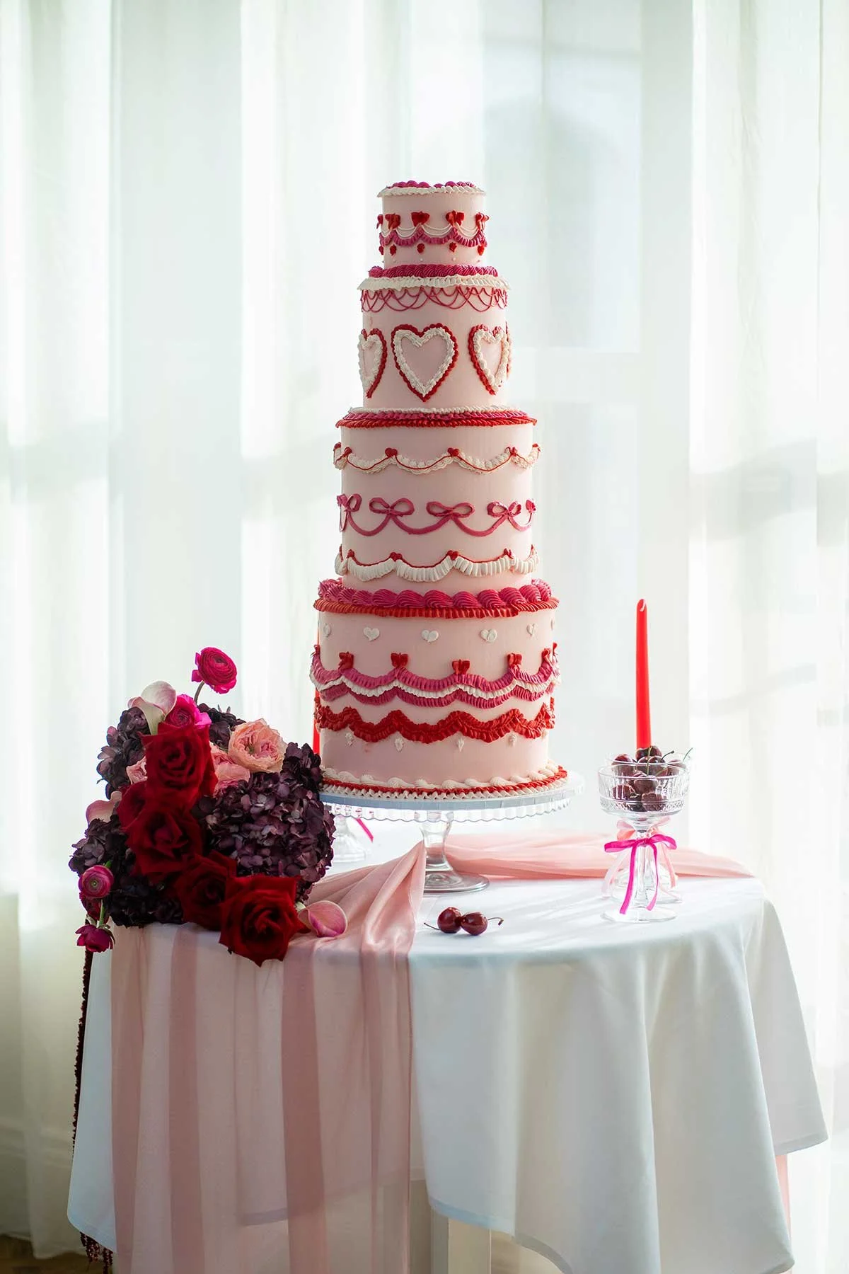 A multi-tiered pink and red Valentine's Day inspired wedding cake on a table with a bouquet of red and pink roses, cherries, and a pink candle with cherries in a glass bowl.