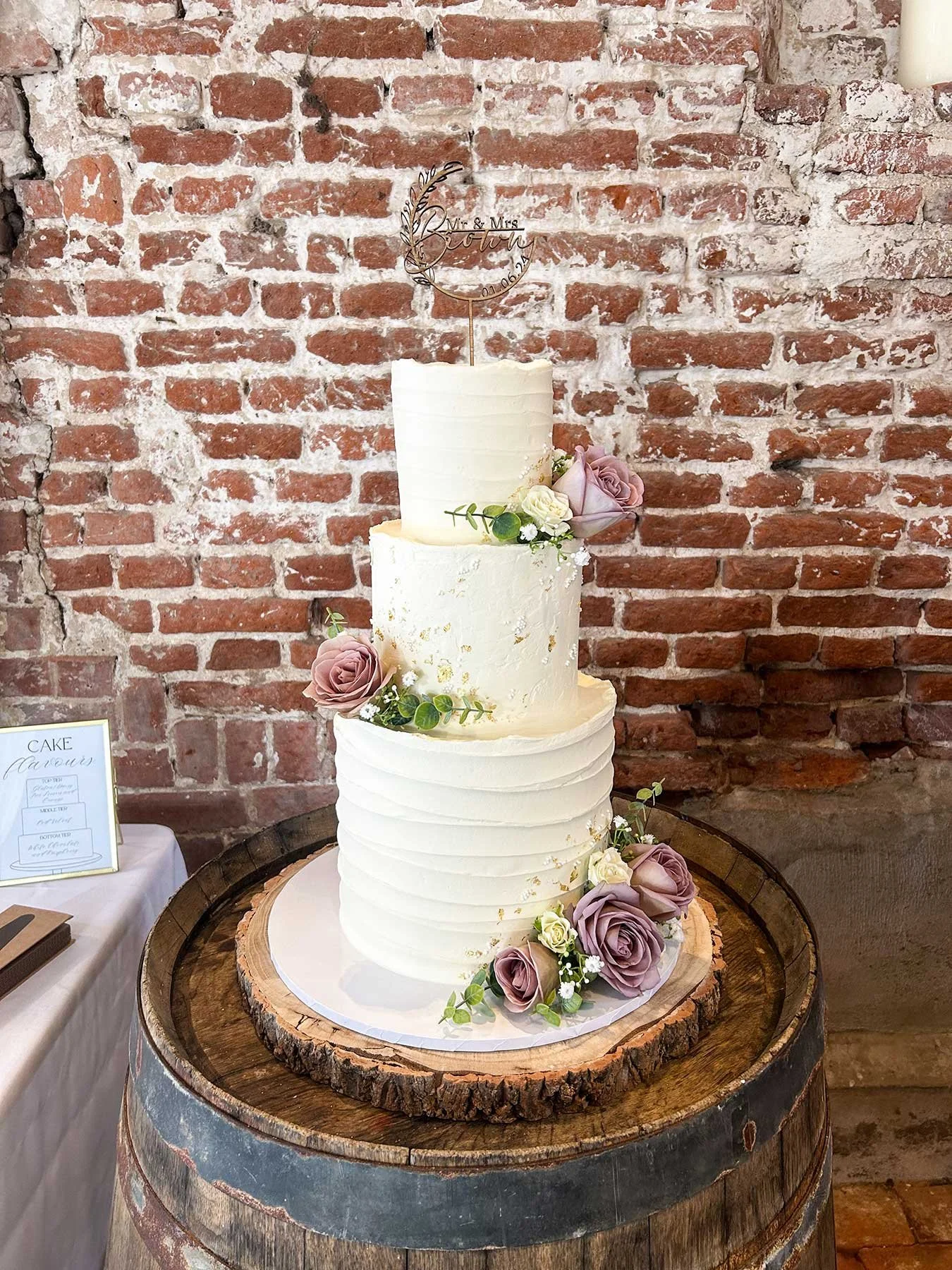 Three-tier white wedding cake with textured horizontal lines, decorated with pink and white roses and green foliage, topped with a metal cake topper reading 'Mr. & Mrs. Brown.'