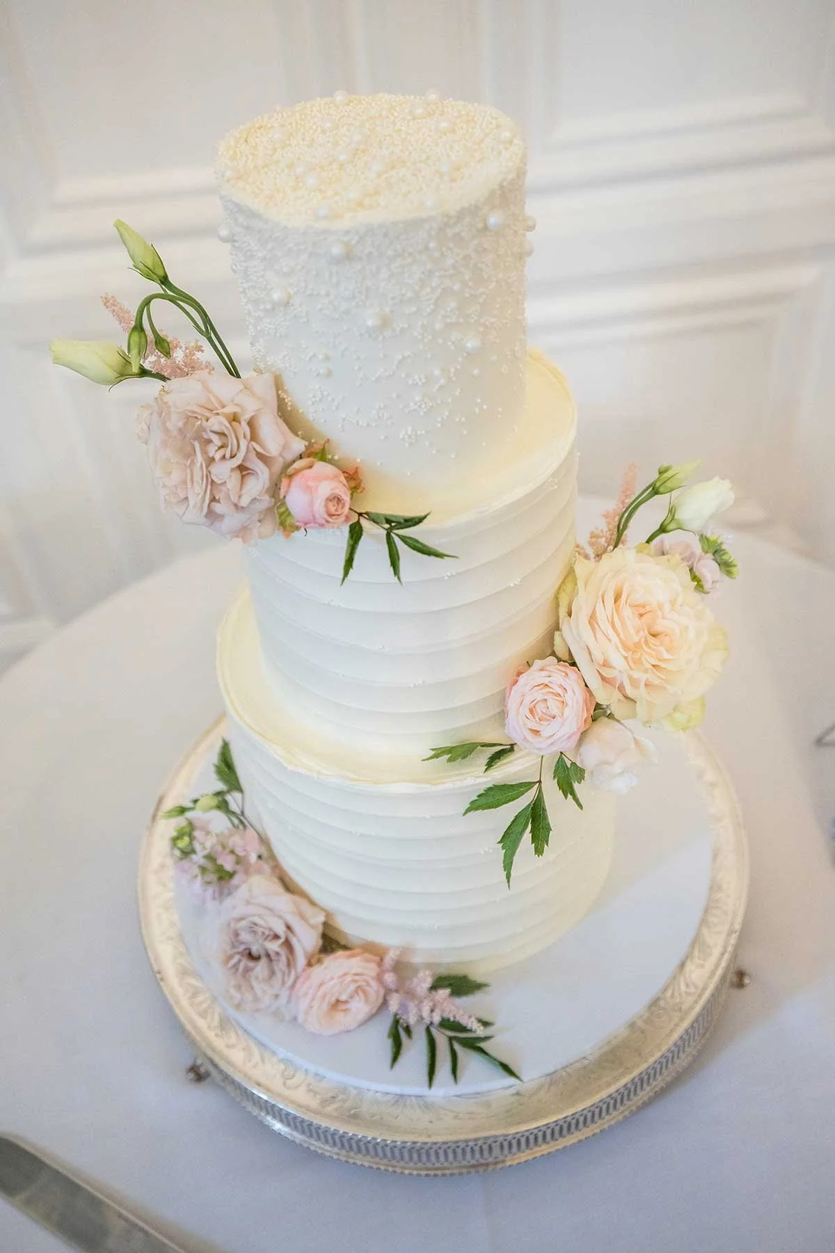 Elegant three-tiered white wedding cake decorated with pastel pink and peach roses, green leaves, and small white pearl-like accents. The cake is positioned on a silver stand with a white tablecloth background.