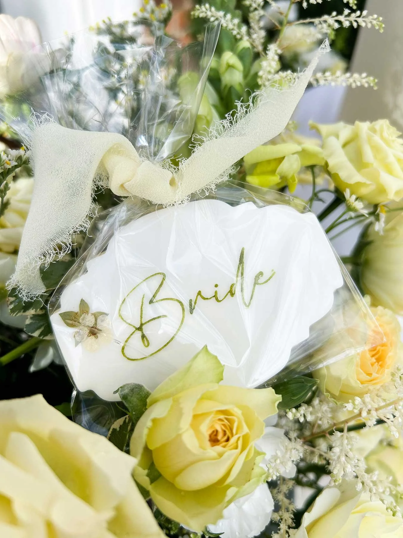 A wedding flower arrangement with a white sign that says 'Bride', surrounded by yellow roses and white flowers, wrapped with a light-colored ribbon.