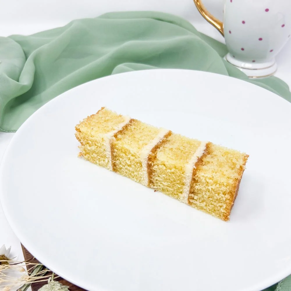A slice of layered yellow cake with white icing on a white plate, with a green cloth and a white mug with purple polka dots in the background.