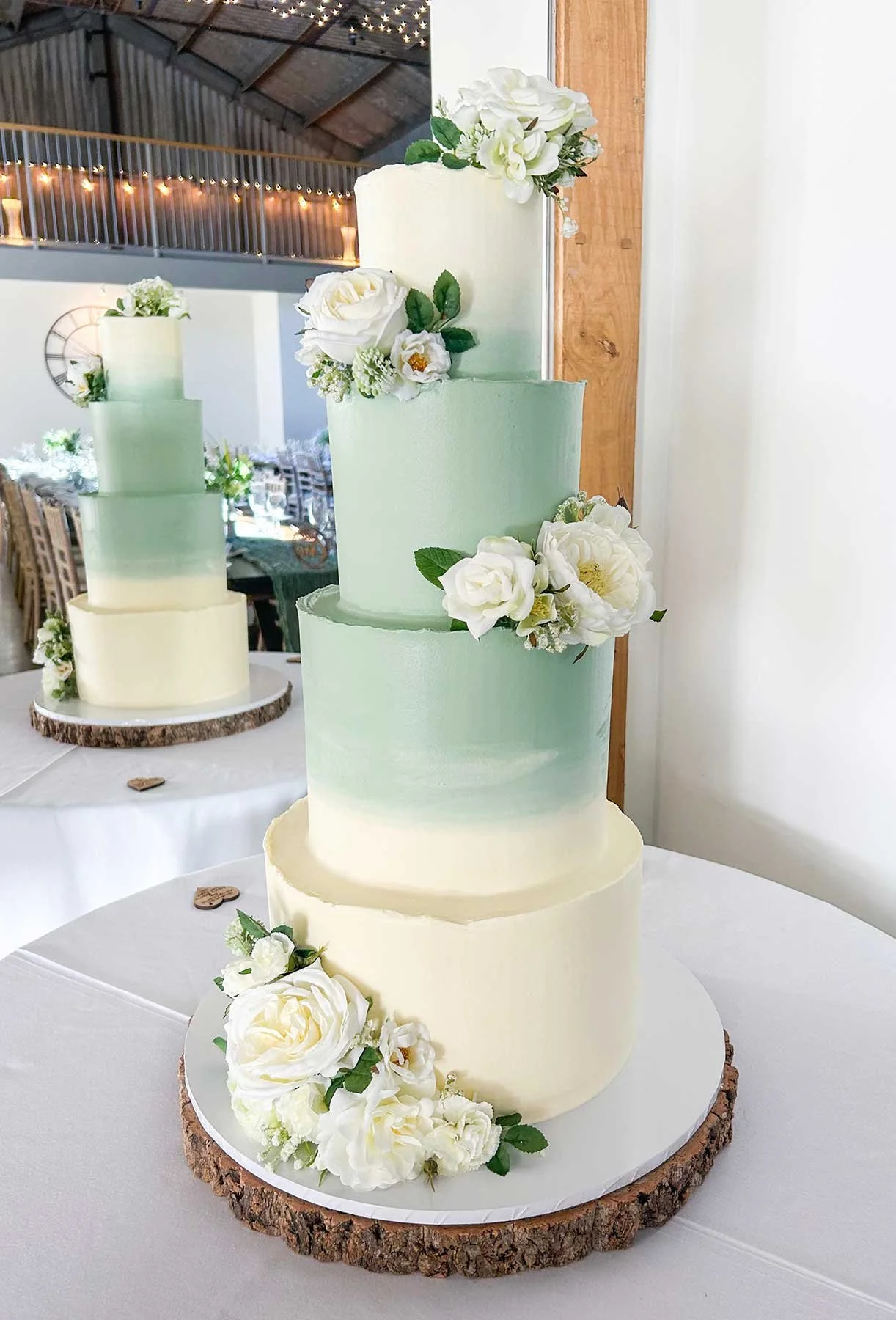A four-tier wedding cake with gradient pastel green and cream frosting, decorated with white roses and greenery, sitting on a wooden round cake base.