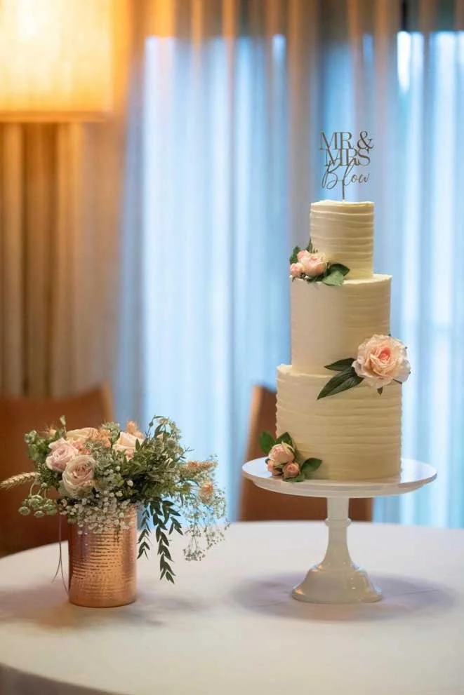 Three-tiered white wedding cake decorated with pink flowers and green leaves, with a 'Mr & Mrs Blow' cake topper, on a white table with a floral arrangement in a copper vase nearby, against a background of blue curtains.