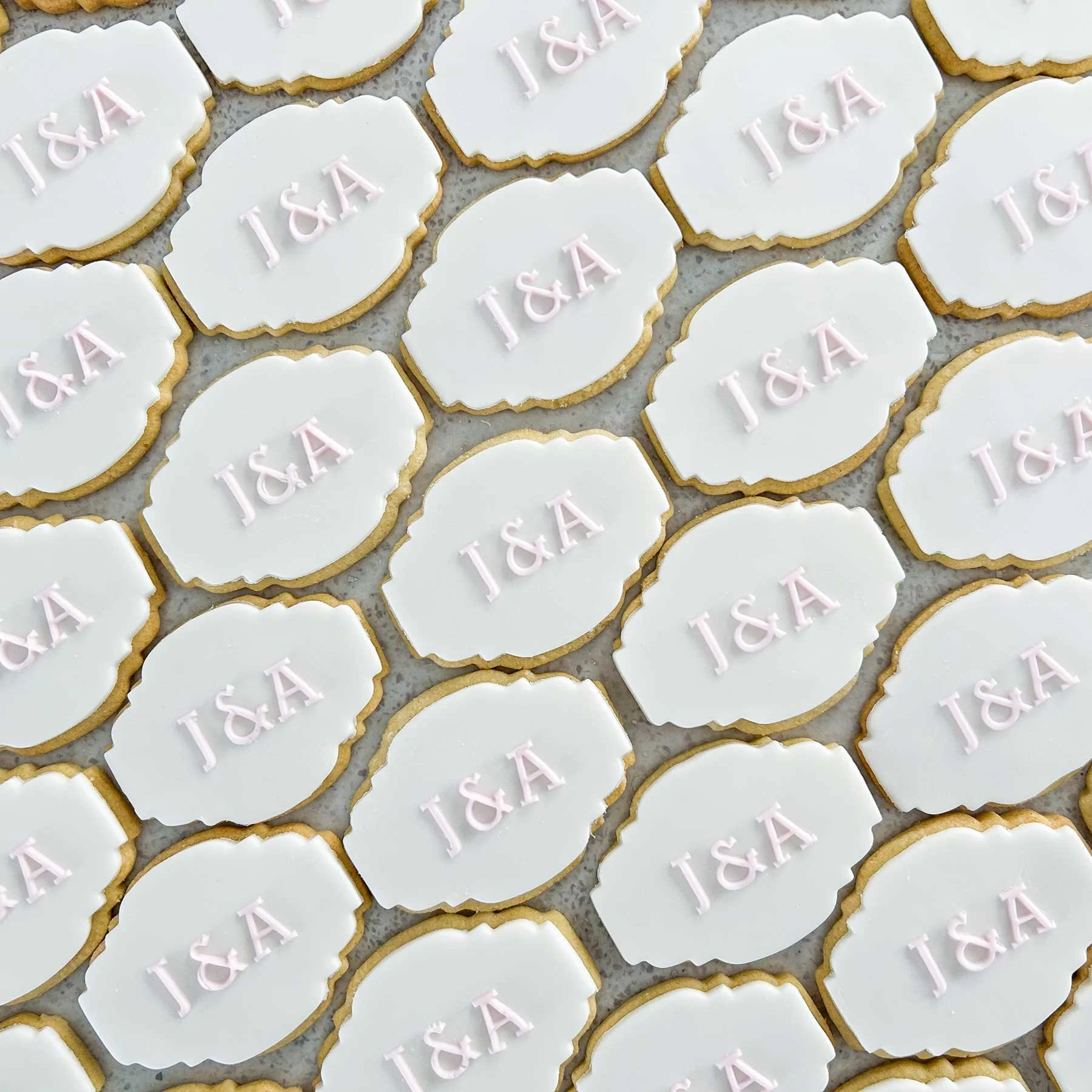Cookies with white icing and pink lettering that spells "J&A" on each cookie, arranged in a pattern on parchment paper.