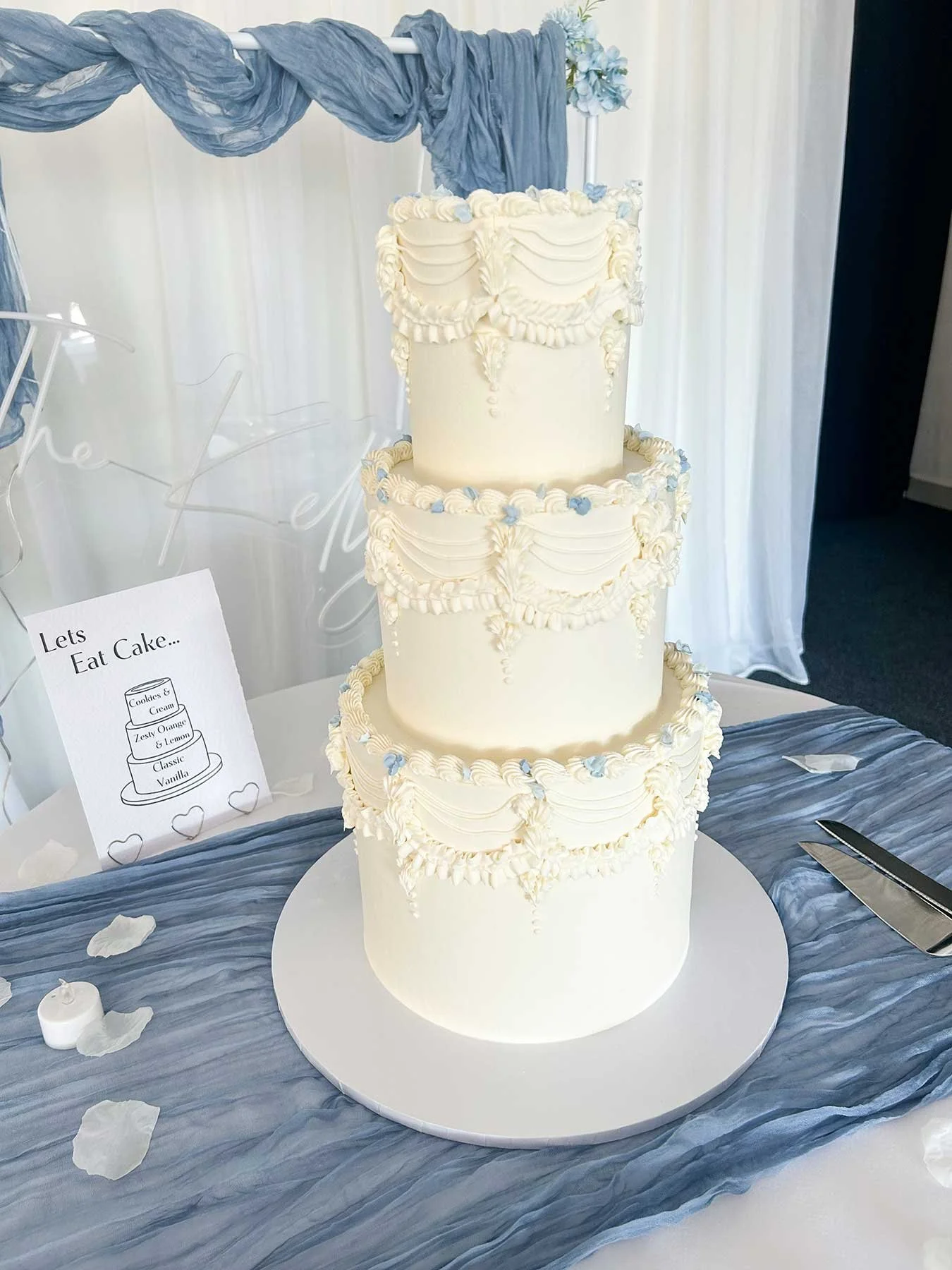 Three-tier white wedding cake with cream frosting and decorative piping, placed on a white cake board on blue fabric, with a sign and scattered white rose petals nearby.