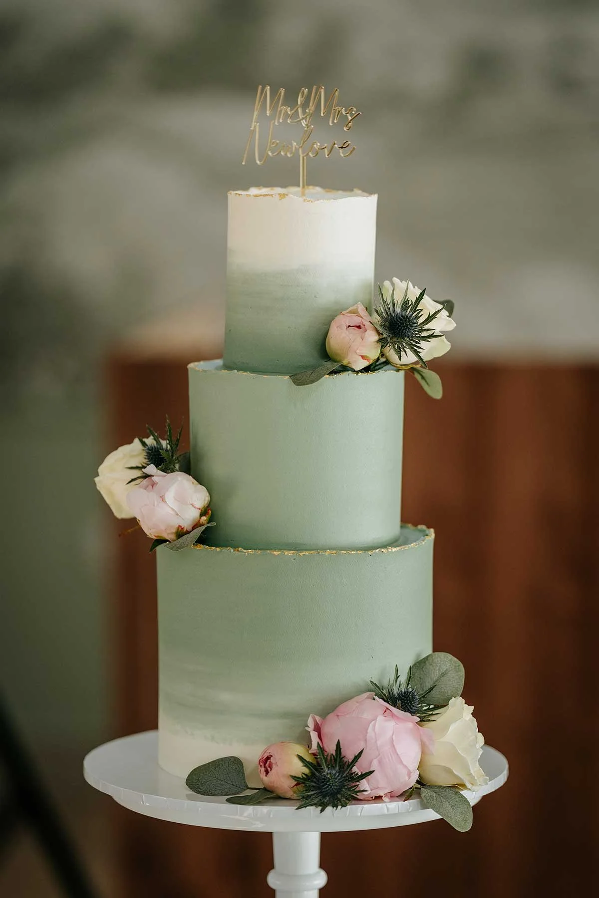 A three-tier wedding cake with a gradient green icing, adorned with pink and white flowers and green leaves. A gold topper reads 'Mr & Mrs Nardore'.