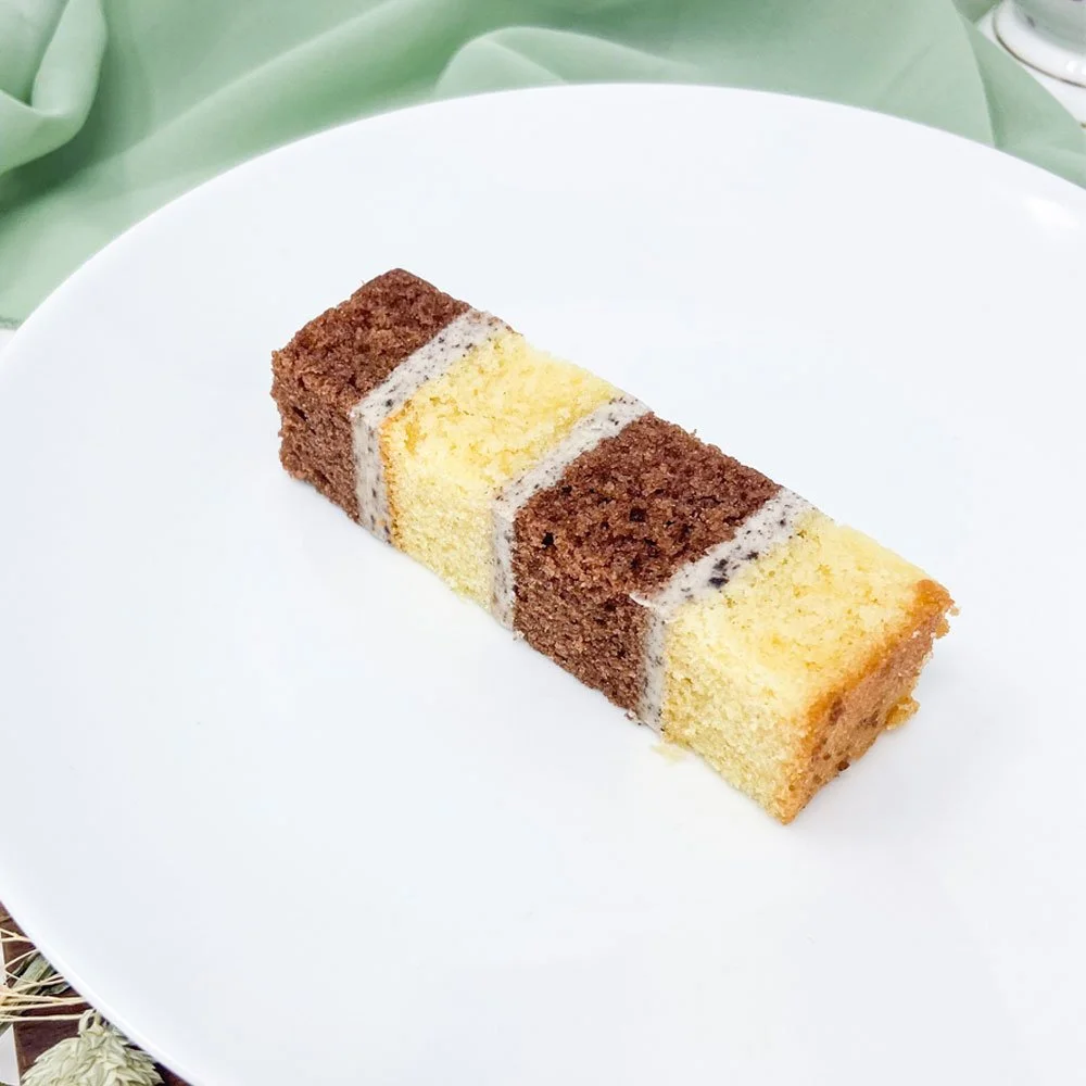 A slice of layered marble cake with yellow and chocolate sponge, topped with white icing, on a white plate.
