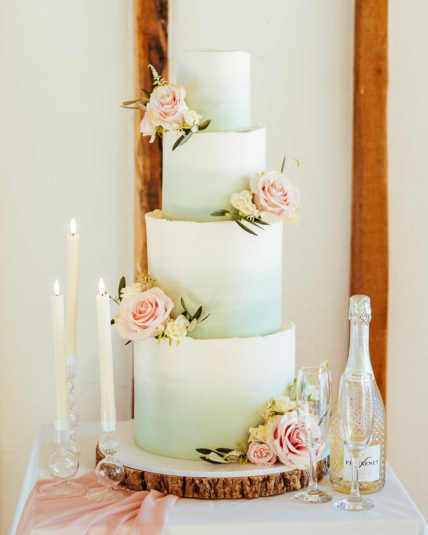 A four-tiered wedding cake decorated with pink and cream roses, placed on a wooden round base, with candles and a champagne bottle on the side.