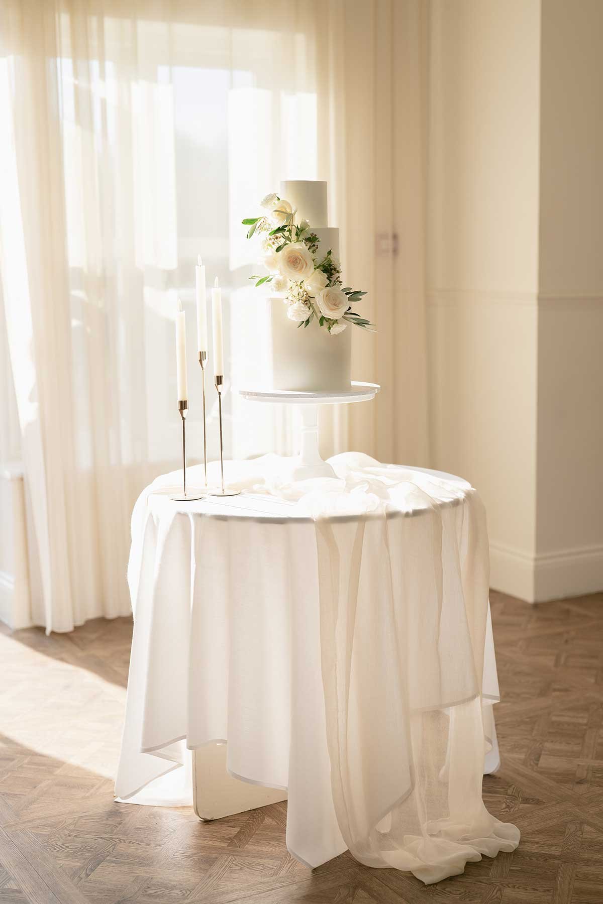 Elegant white wedding cake with floral decoration and four white candles on a round table, in a softly lit room.