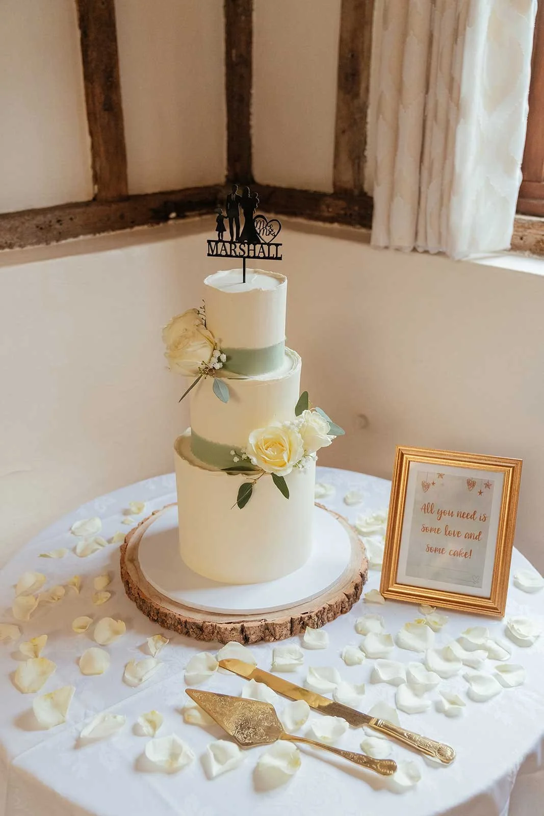 Three-tier white wedding cake decorated with white roses and greenery, placed on a wooden slab, surrounded by white flower petals and gold cake serving utensils on a table. A framed sign reads, 'All you need is some love and some cake!'