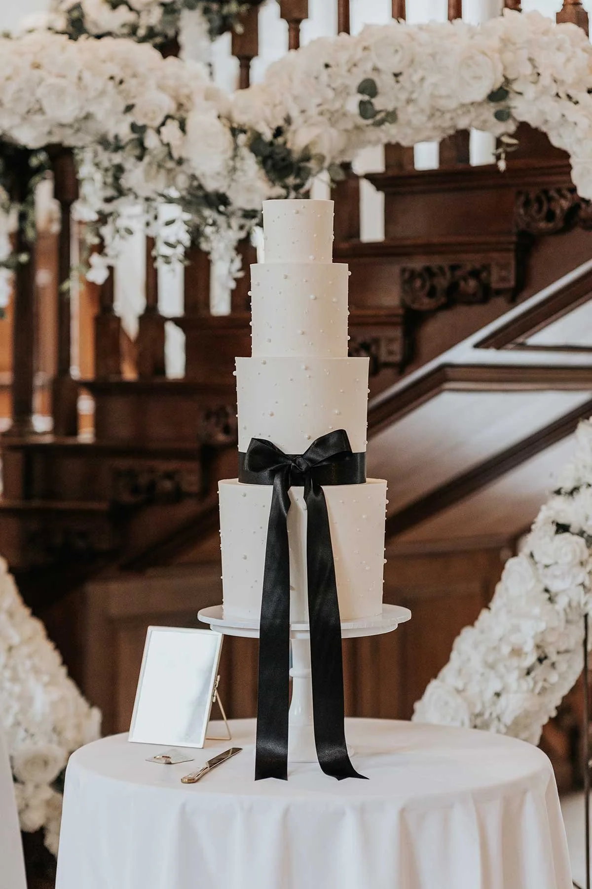 A tall, white four-tier wedding cake with a black ribbon tied in a bow on the front, displayed on a white round table in a decorated venue with floral arrangements and wooden stair railing in the background.