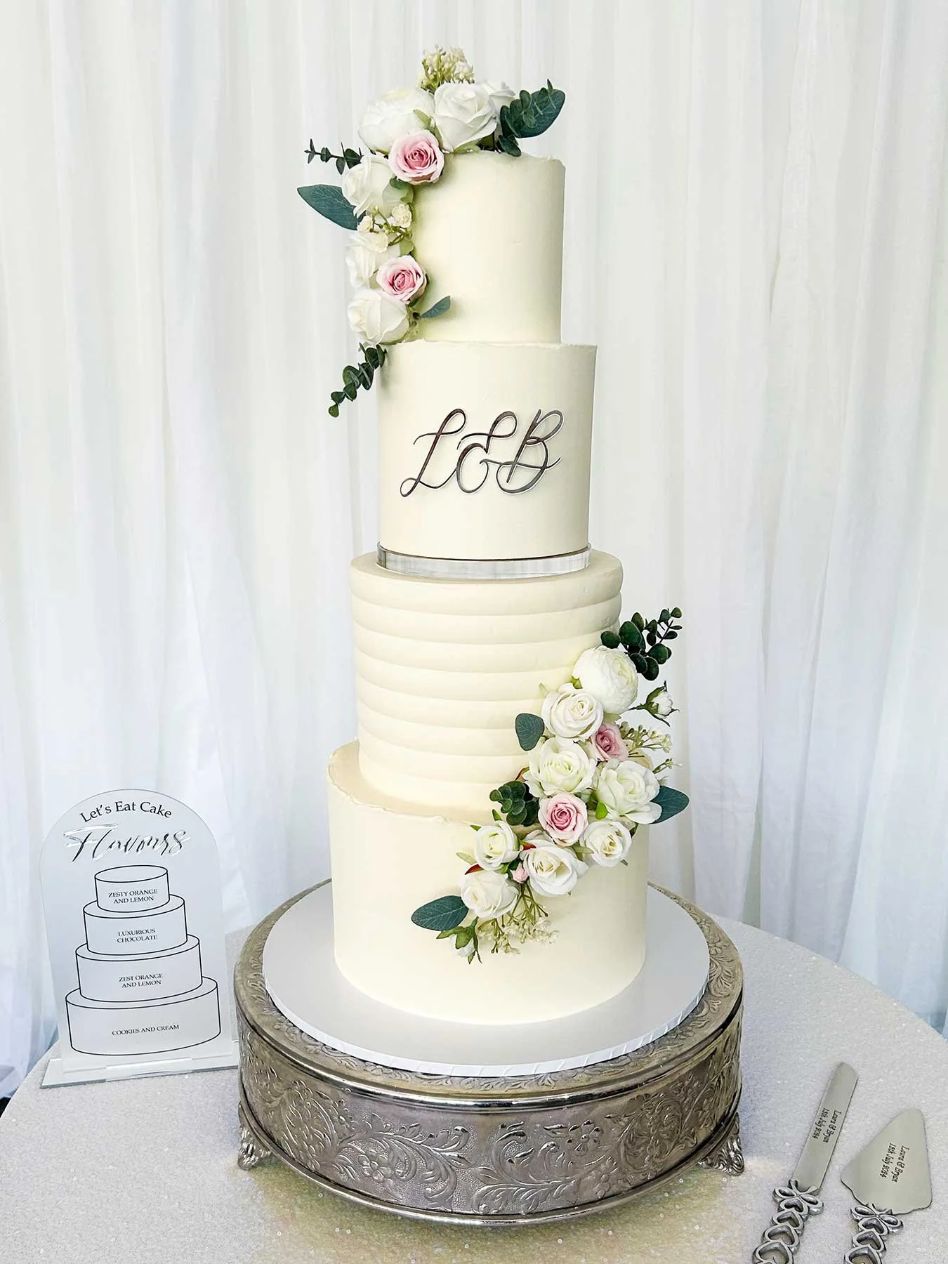 A four-tier wedding cake decorated with white and pink roses and green foliage, featuring monogram initials 'L & B' on the second tier, displayed on a silver ornate cake stand with a white background.