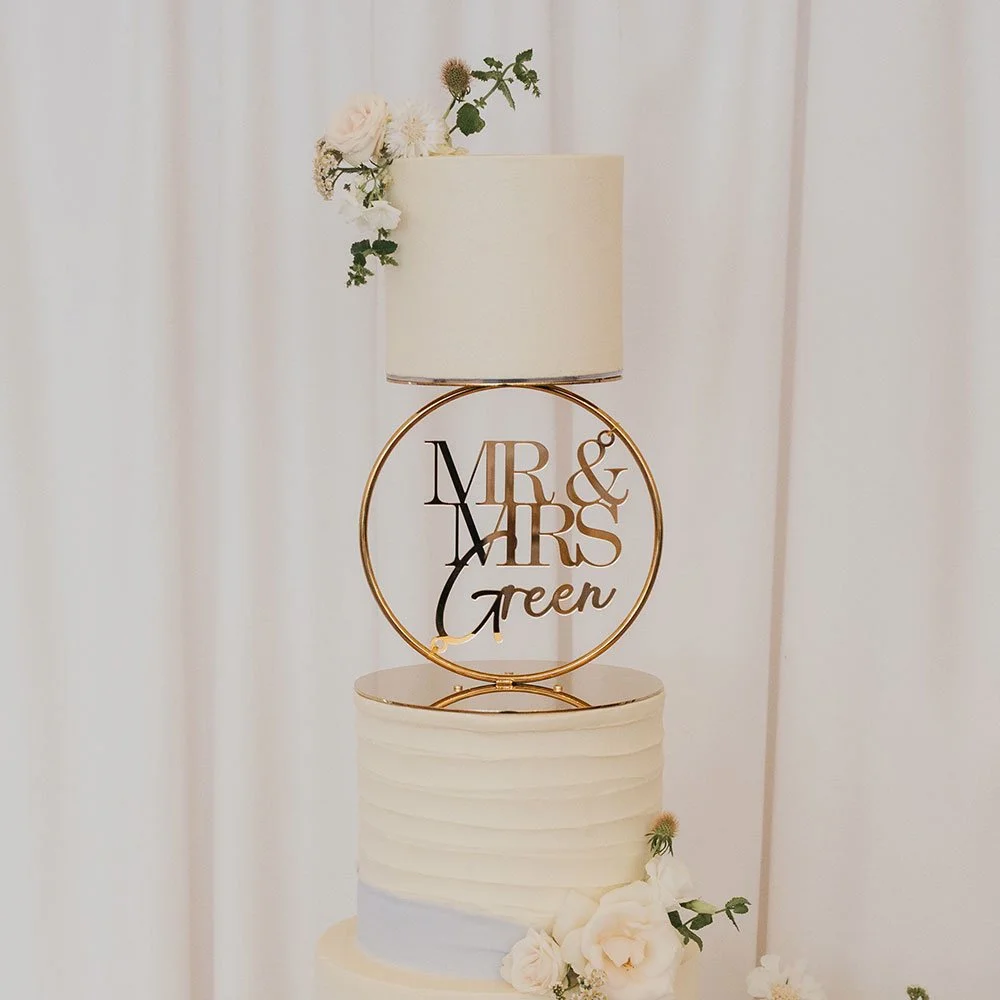 A wedding cake decorated with white flowers and a gold topper that reads 'Mr & Mrs Green'. The cake is white with horizontal ridges, and above it is a table lamp with a white shade, topped with flowers.
