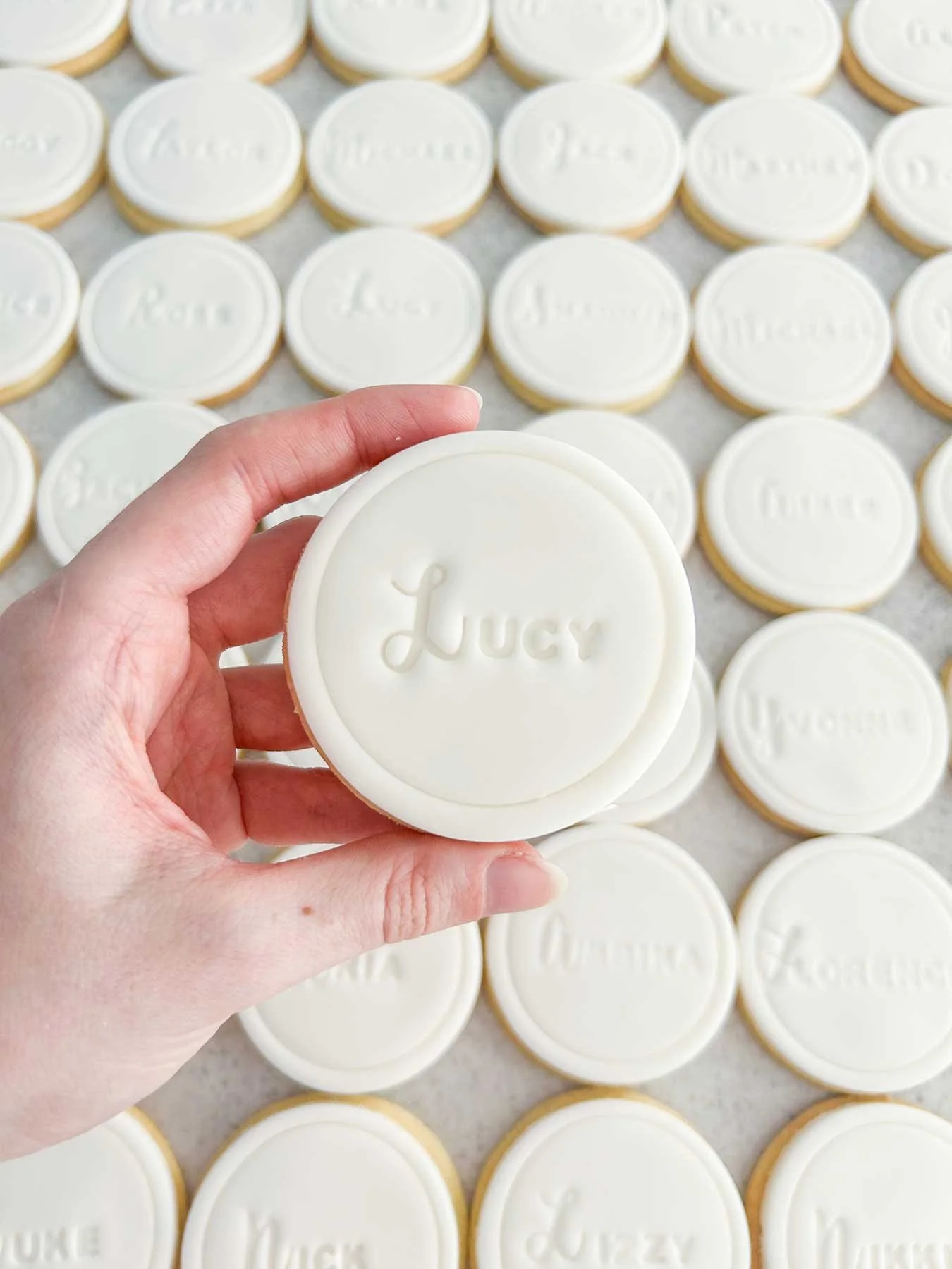 A hand holding a round sugar cookie with white icing, embossed with the name 'Lucy'. Several similar cookies with the same embossed name are laid out on a surface in the background.