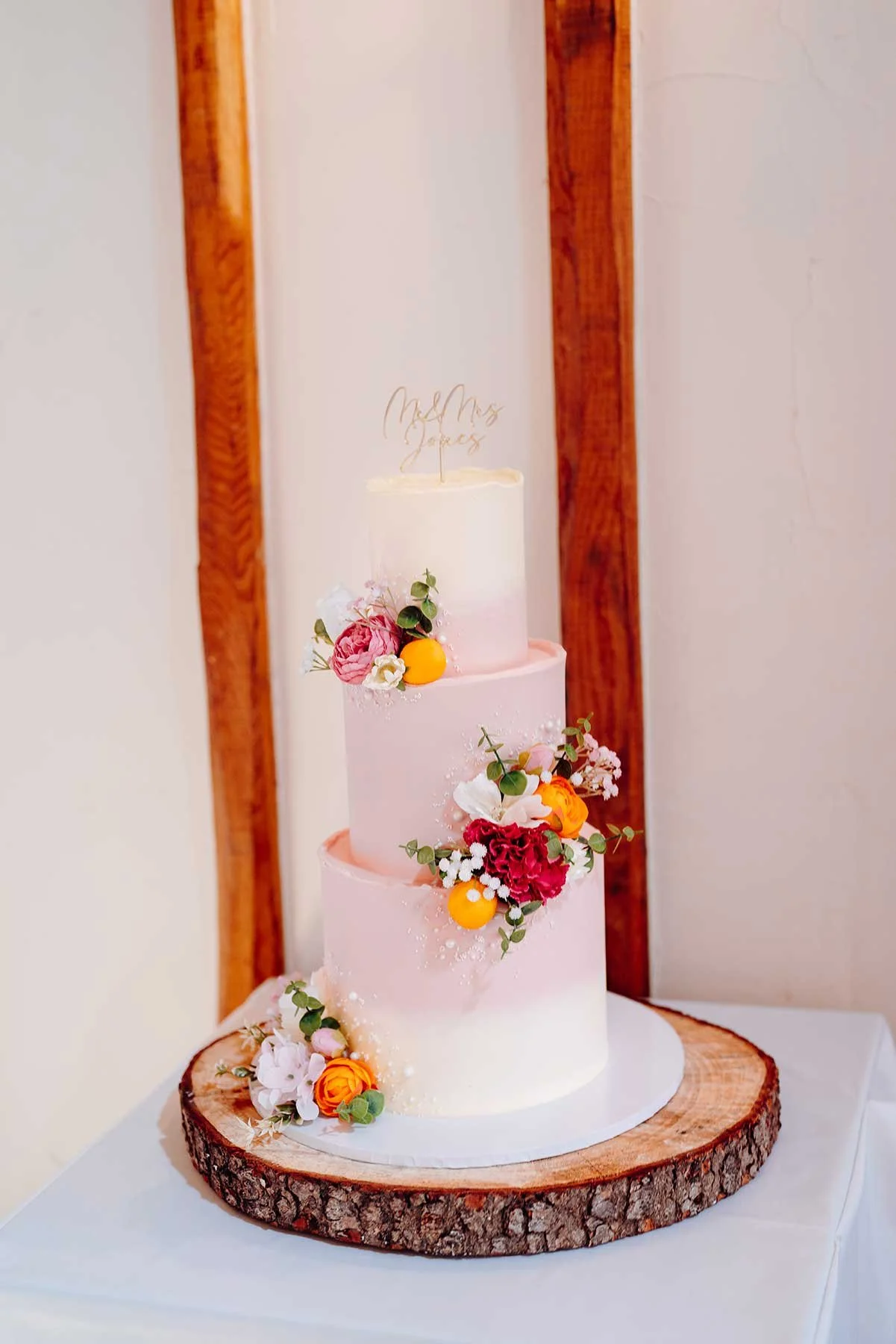 A three-tiered wedding cake with pink and white gradient frosting, decorated with colorful flowers and small fruit accents, sitting on a wooden slab.
