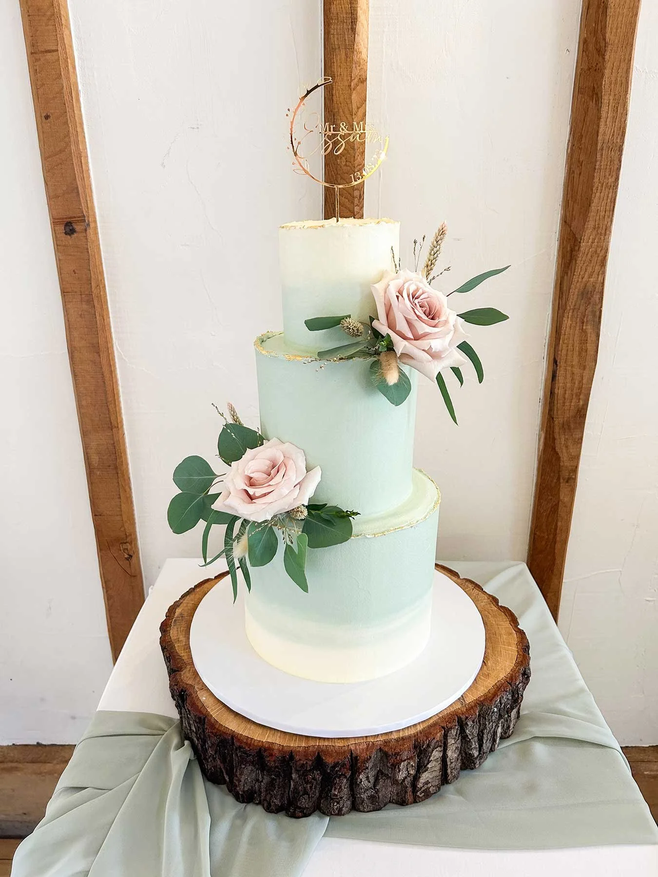 A three-tier wedding cake with a rustic wood base, decorated with blush pink roses and greenery, topped with a gold crescent moon cake topper that says 'Mr & Mrs Essam' and a date; the cake has subtle pastel shades and gold accents.