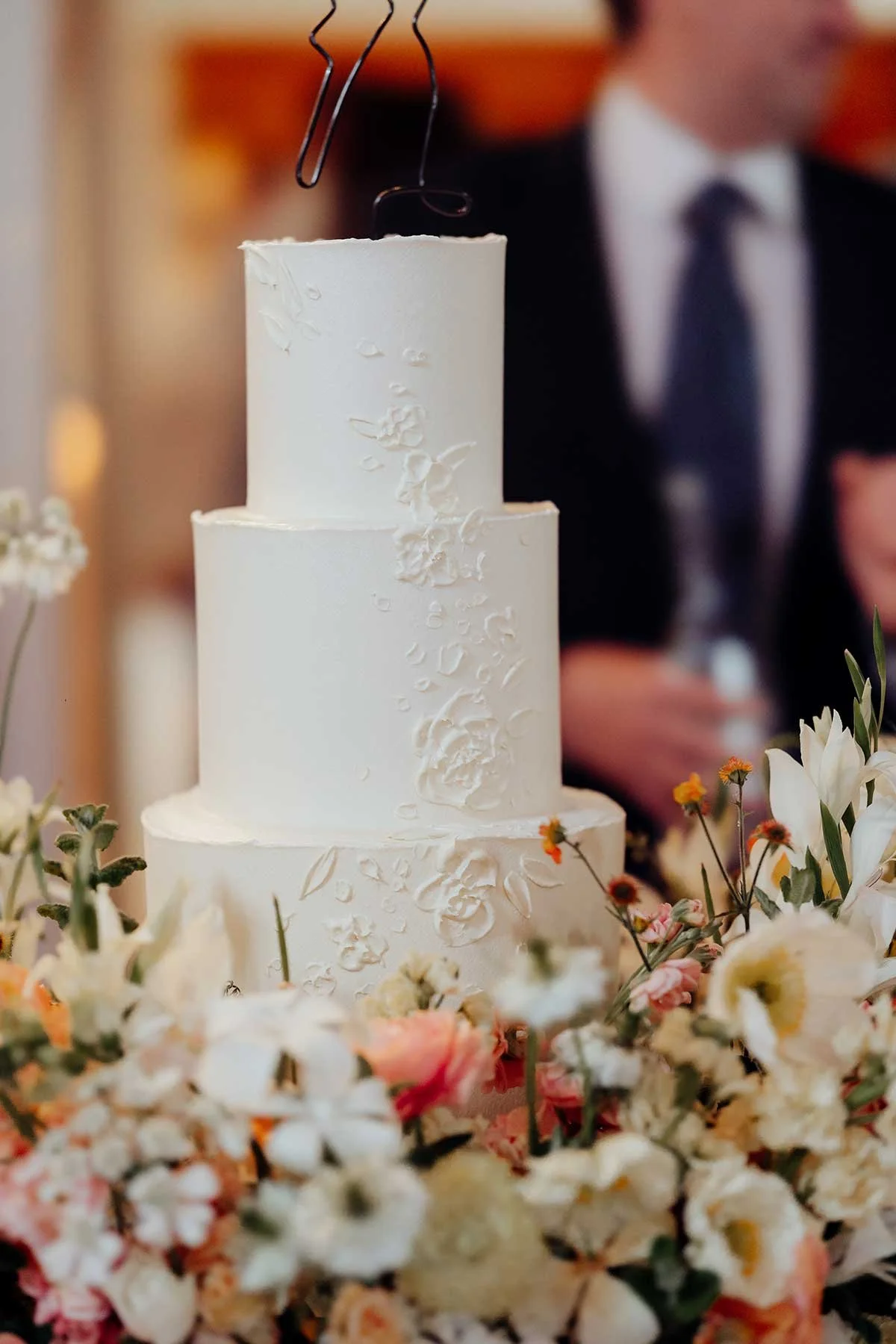 Three-tiered white wedding cake with textured floral design, decorated with colorful flowers around the base, with a person in formal attire blurred in the background.